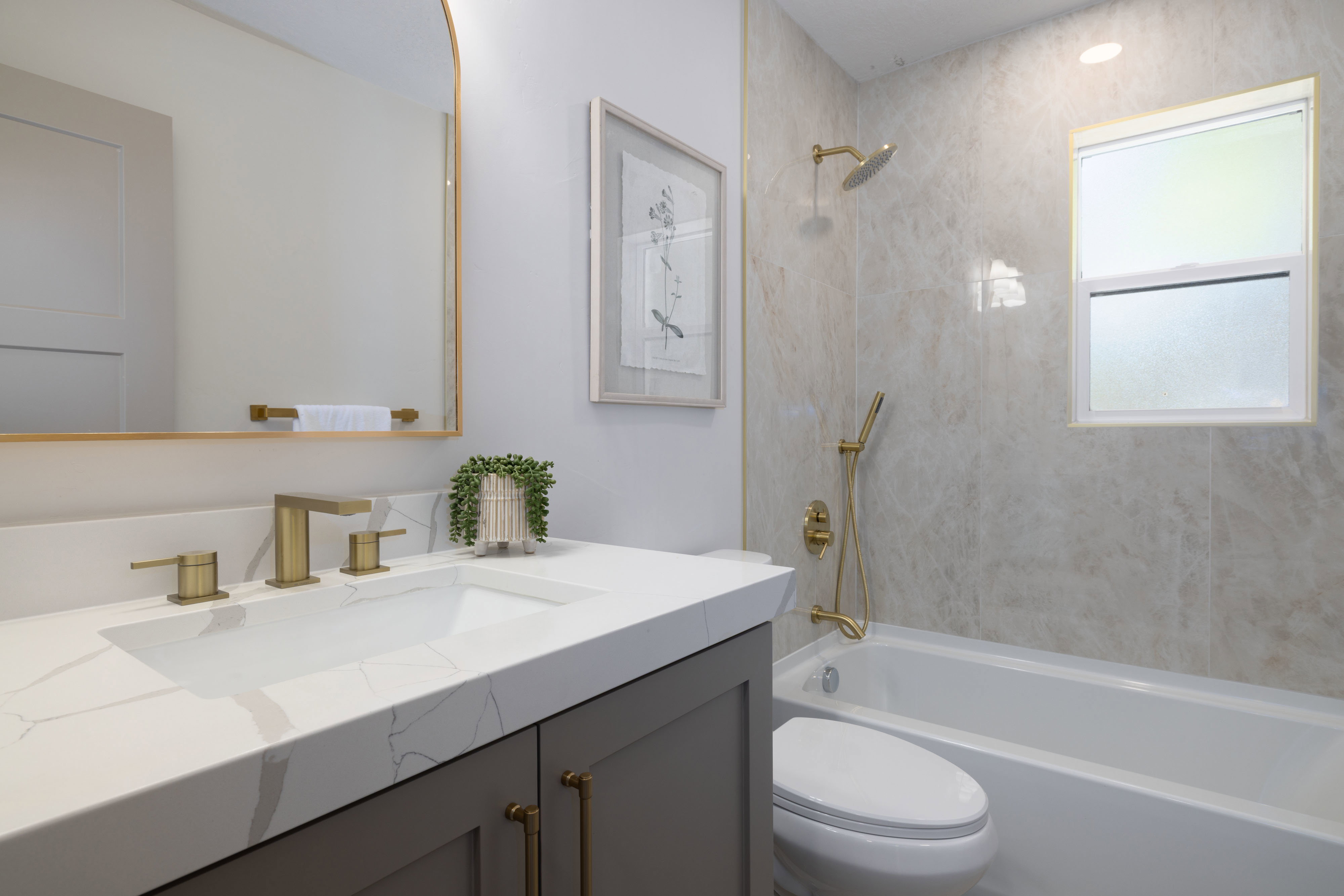 Bathroom featuring a white bathtub with gold-finish fixtures, a window, and large format wall tiling