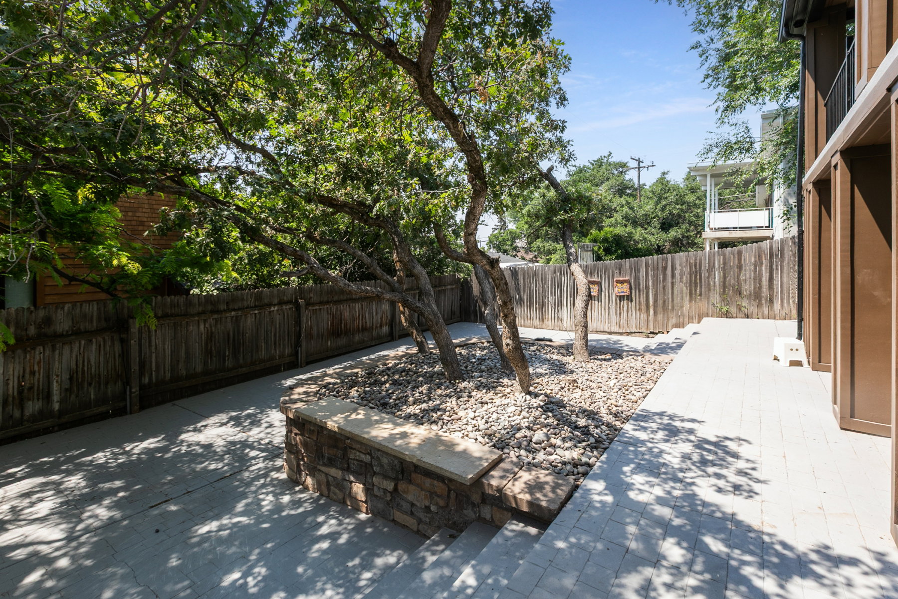 Outdoor patio area featuring light-colored pavers, mature trees, and a stone-clad raised planter with stone steps