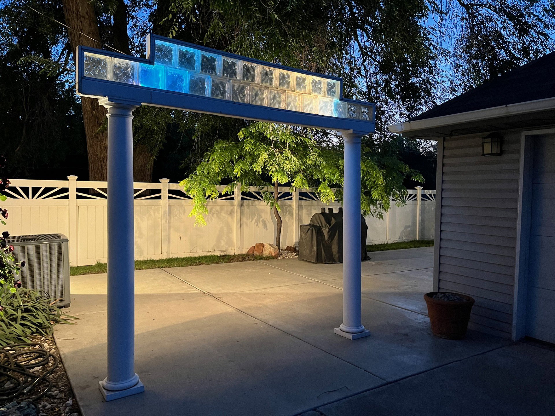 Outdoor covered patio area featuring a decorative archway with glass block accents, smooth concrete flooring, and a white privacy fence