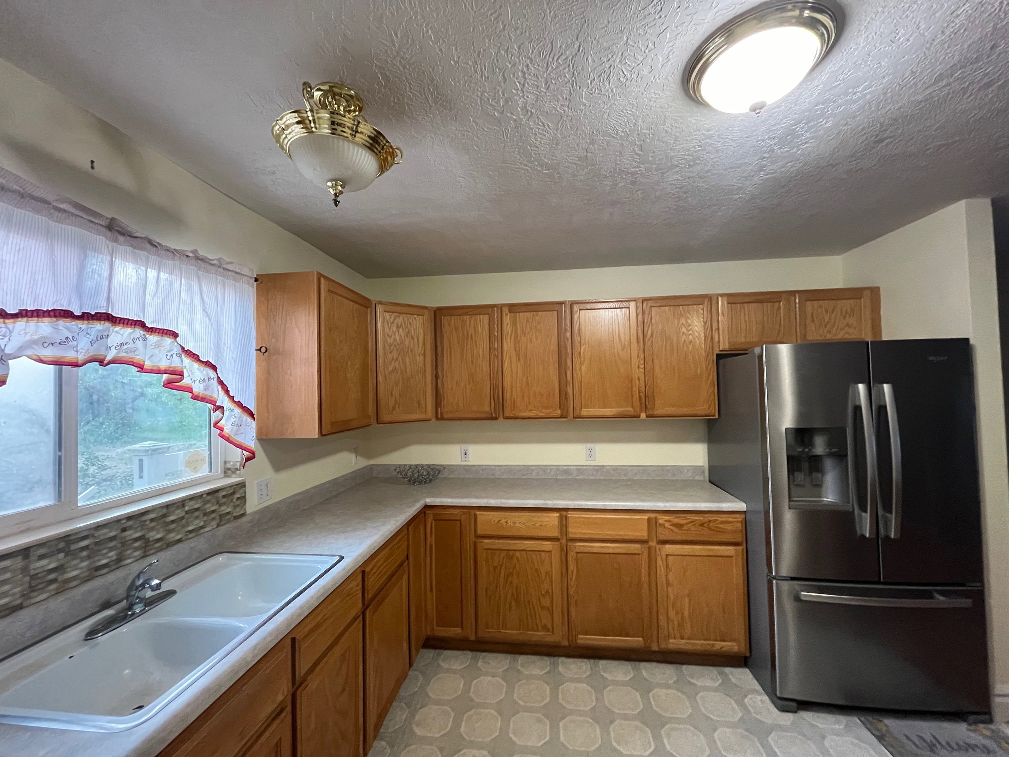 Kitchen featuring wood cabinetry, L-shaped countertops, a double basin sink with a chrome faucet, and a stainless steel refrigerator