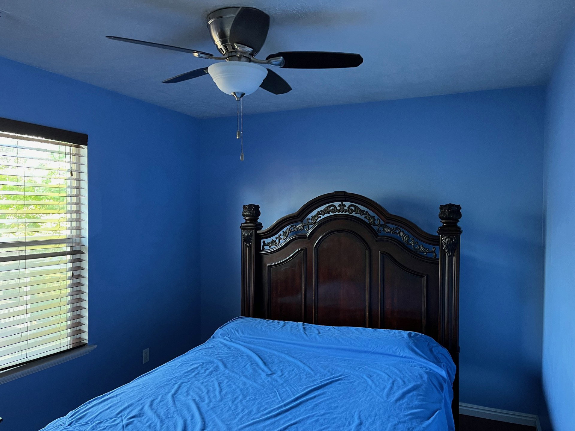 Room with blue walls featuring a ceiling fan with light kit, a window with horizontal blinds, and a dark wood-finish headboard