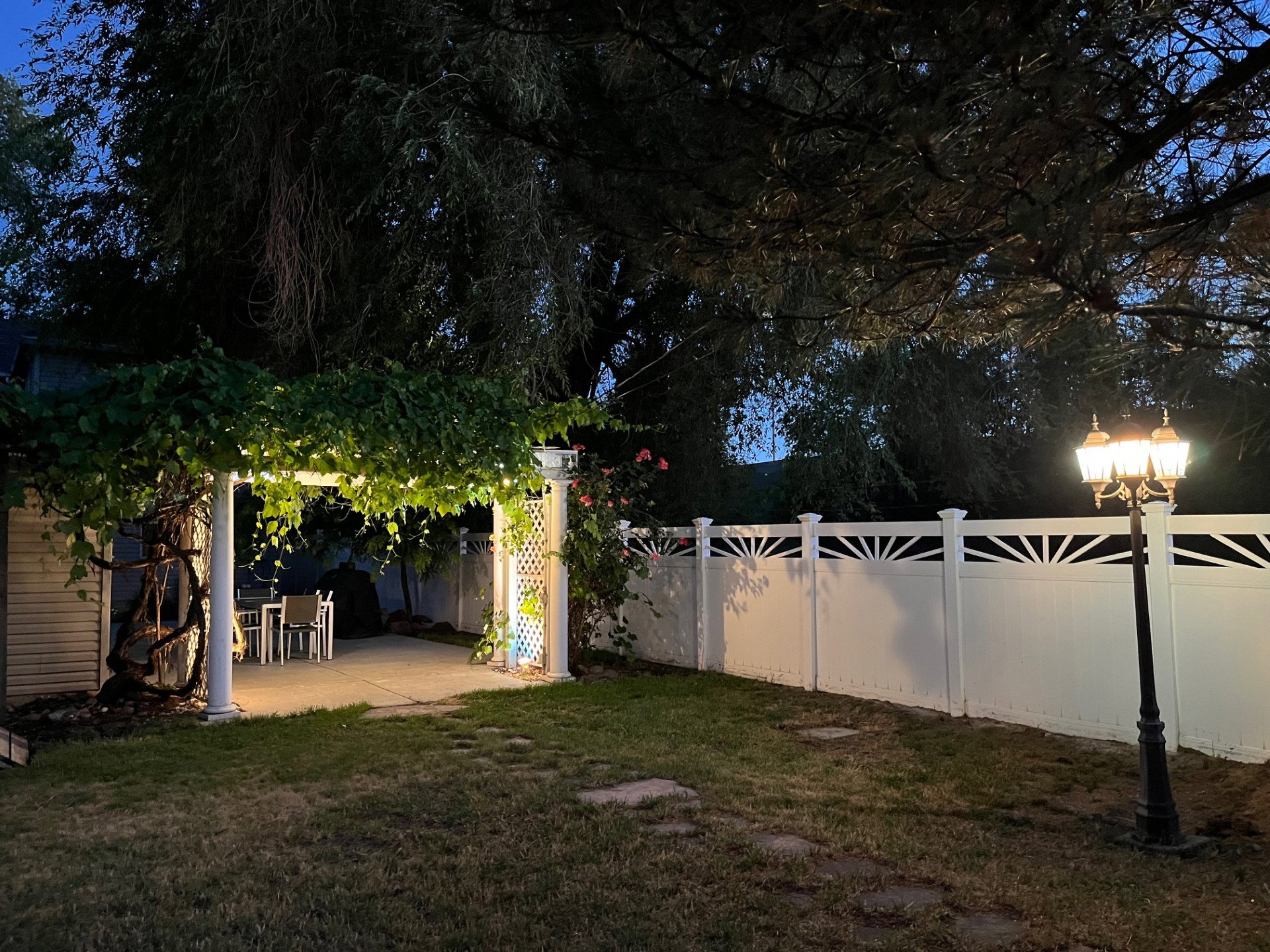 Backyard featuring a covered patio with string lights, a white privacy fence with decorative toppers, and a classic black lamppost