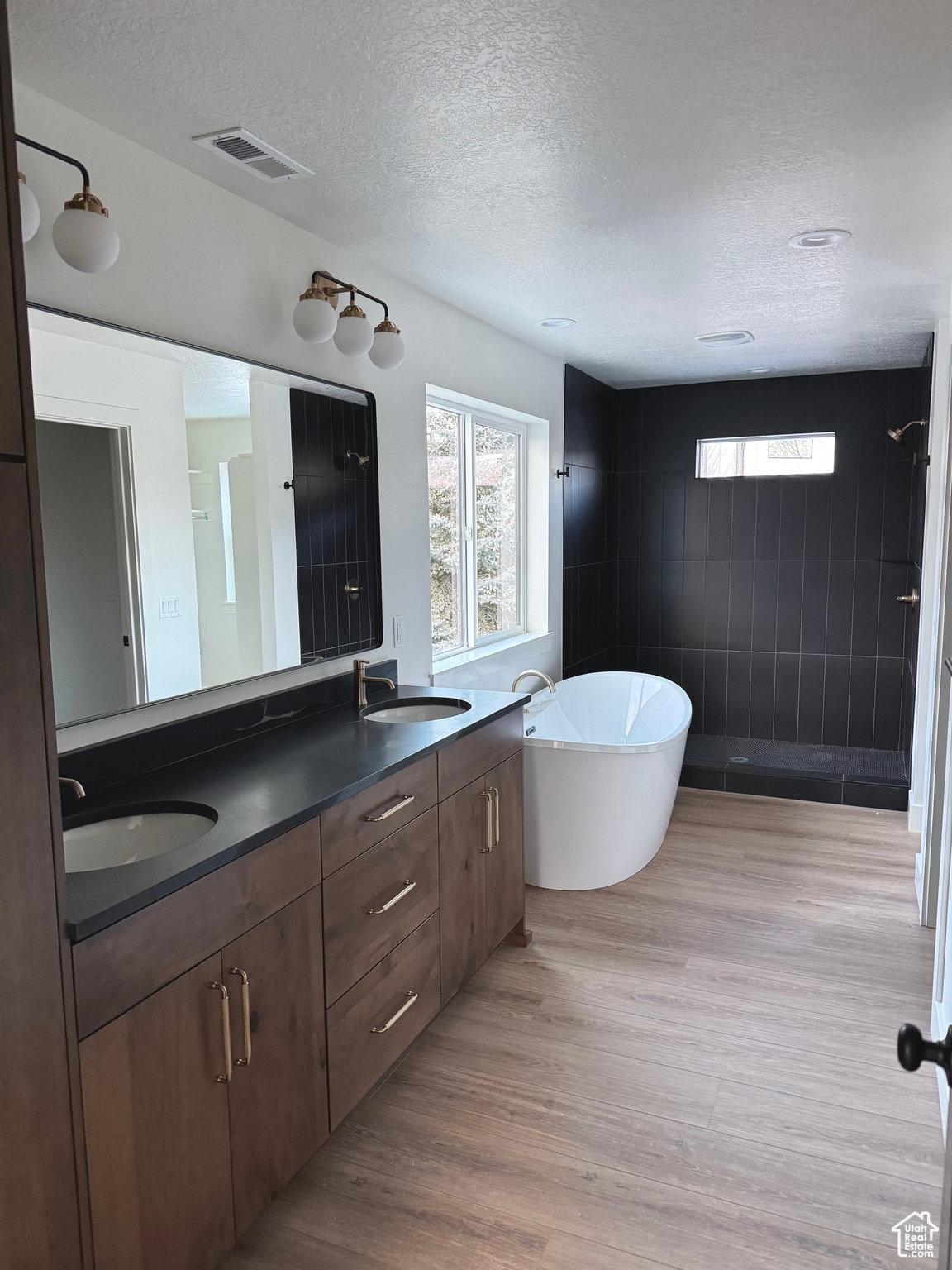 Spacious bathroom featuring a dual vanity with a dark countertop and wood-finish cabinetry, a freestanding soaking tub, a walk-in shower with black subway tile, and wood-finish flooring