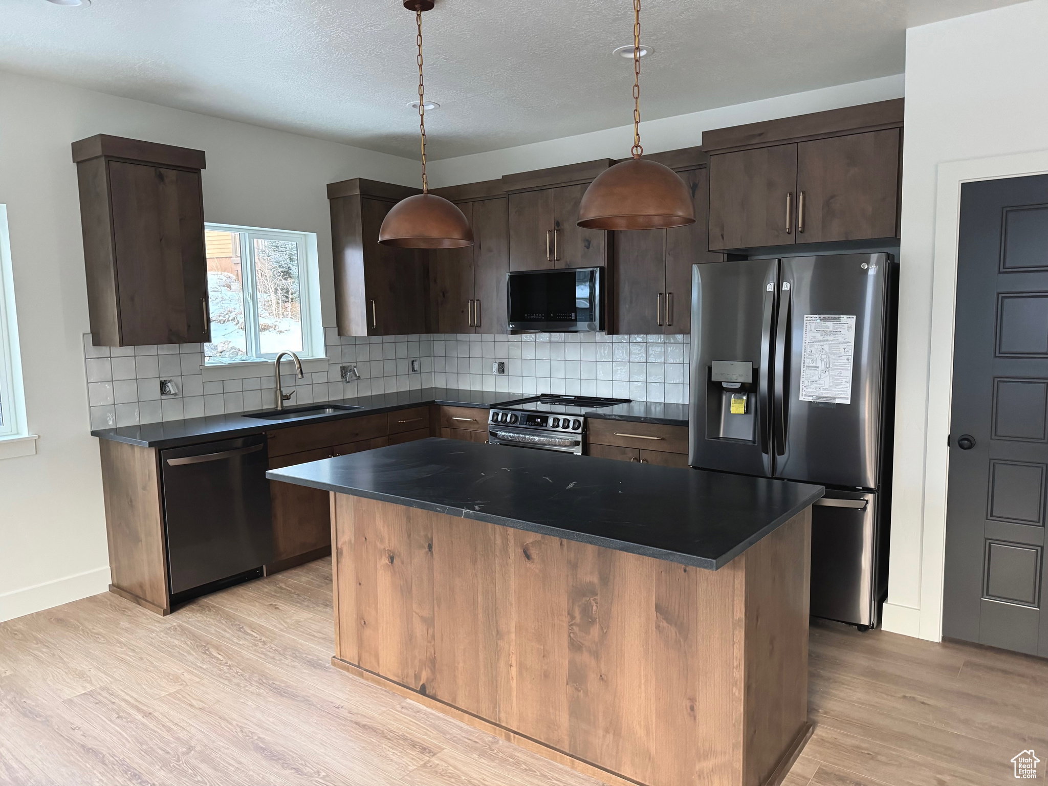 Contemporary kitchen featuring wood-finish cabinetry, stainless steel appliances, dark countertops, a white tiled backsplash, and light wood-finish flooring