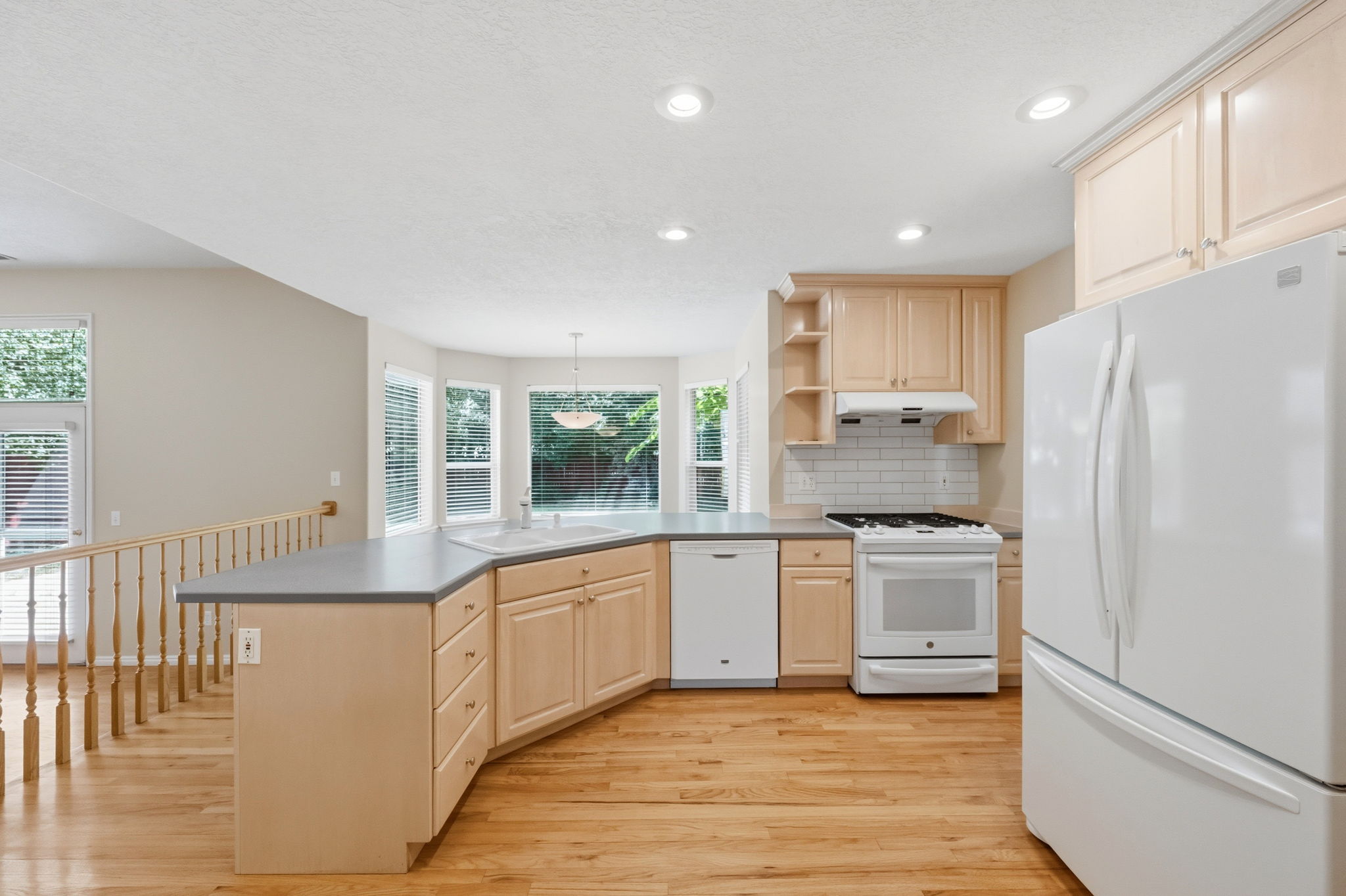 Kitchen featuring wood-finish flooring, light wood cabinetry, a white subway tile backsplash, and a corner island with a double basin sink
