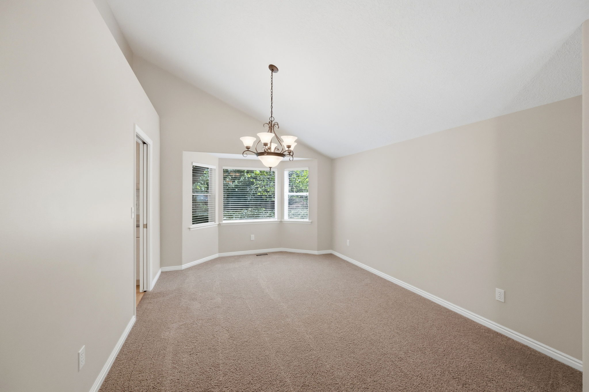 Vaulted ceiling with an ornate chandelier, bay window with blinds, neutral-toned walls, and carpet flooring