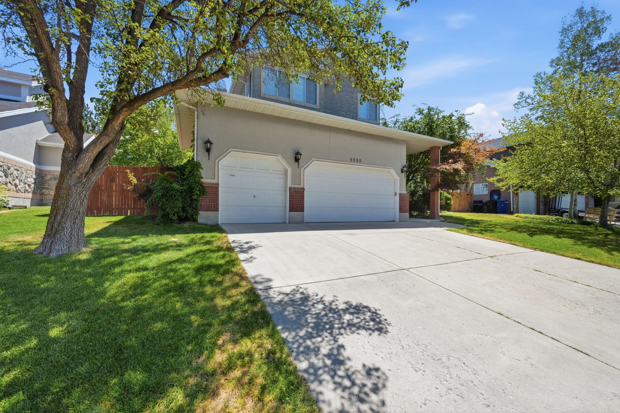 Two-story stucco and brick facade property featuring a two-car garage with white doors, a concrete driveway, and mature landscaping with established trees and green lawn