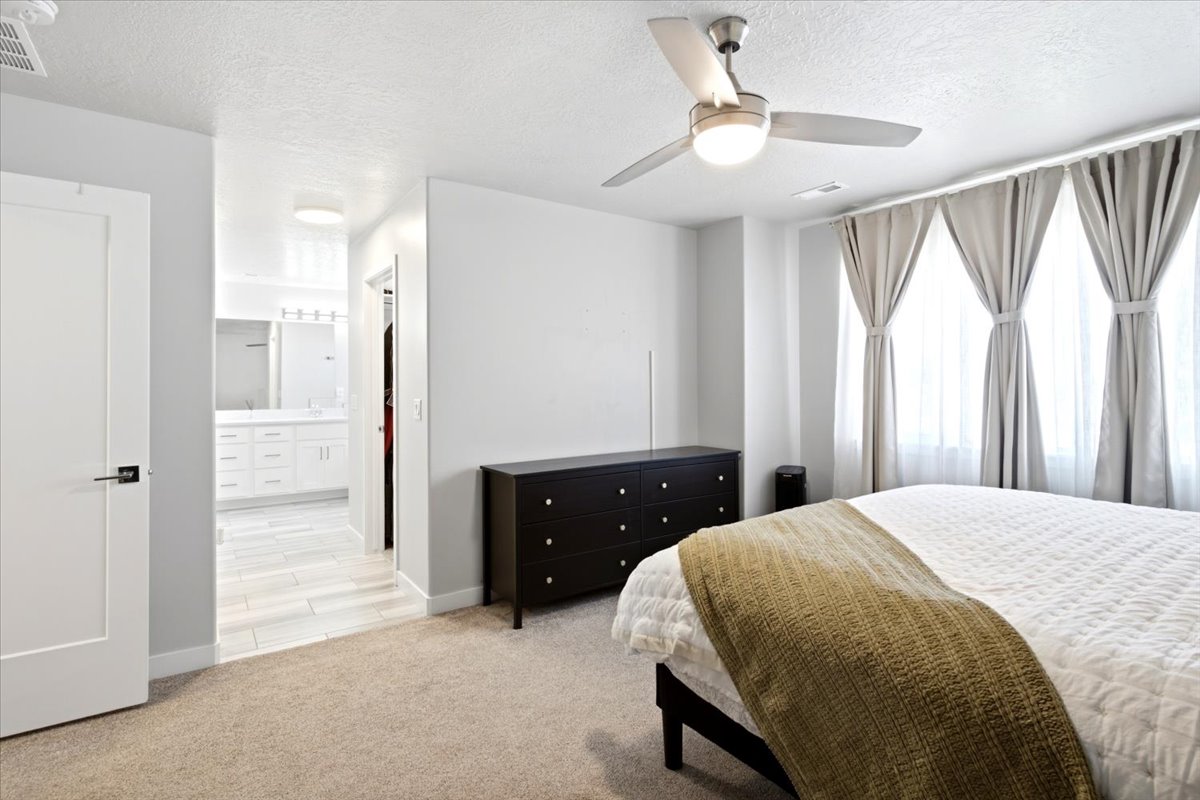 Carpeted bedroom featuring a contemporary ceiling fan with integrated lighting, three large windows, a white paneled door with black hardware, and a clear sightline into an adjacent bathroom with a double vanity