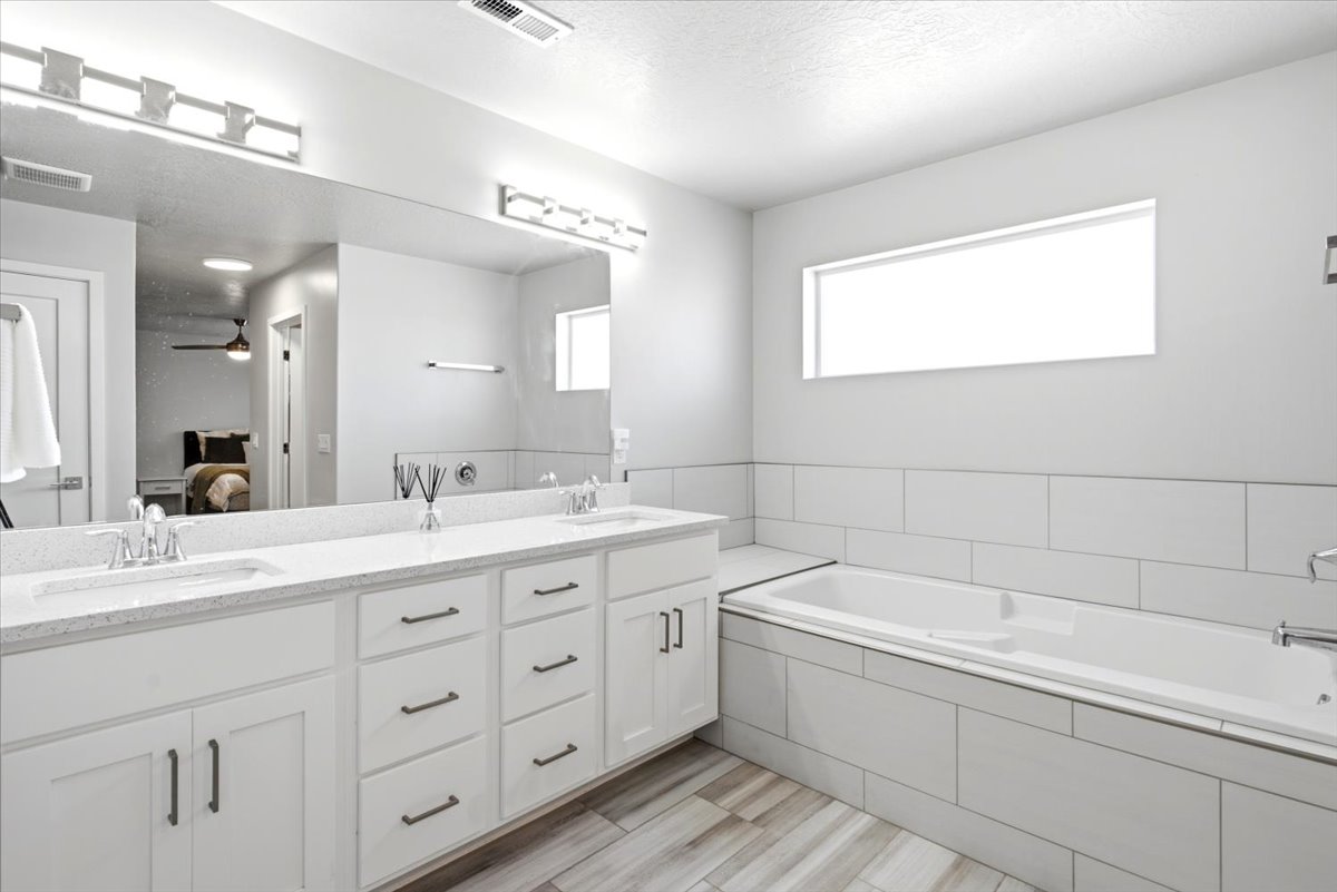 Dual vanity with undermount sinks and light-toned countertops, large mirror, white cabinetry with dark hardware, built-in bathtub with tiled surround, and wood-finish flooring