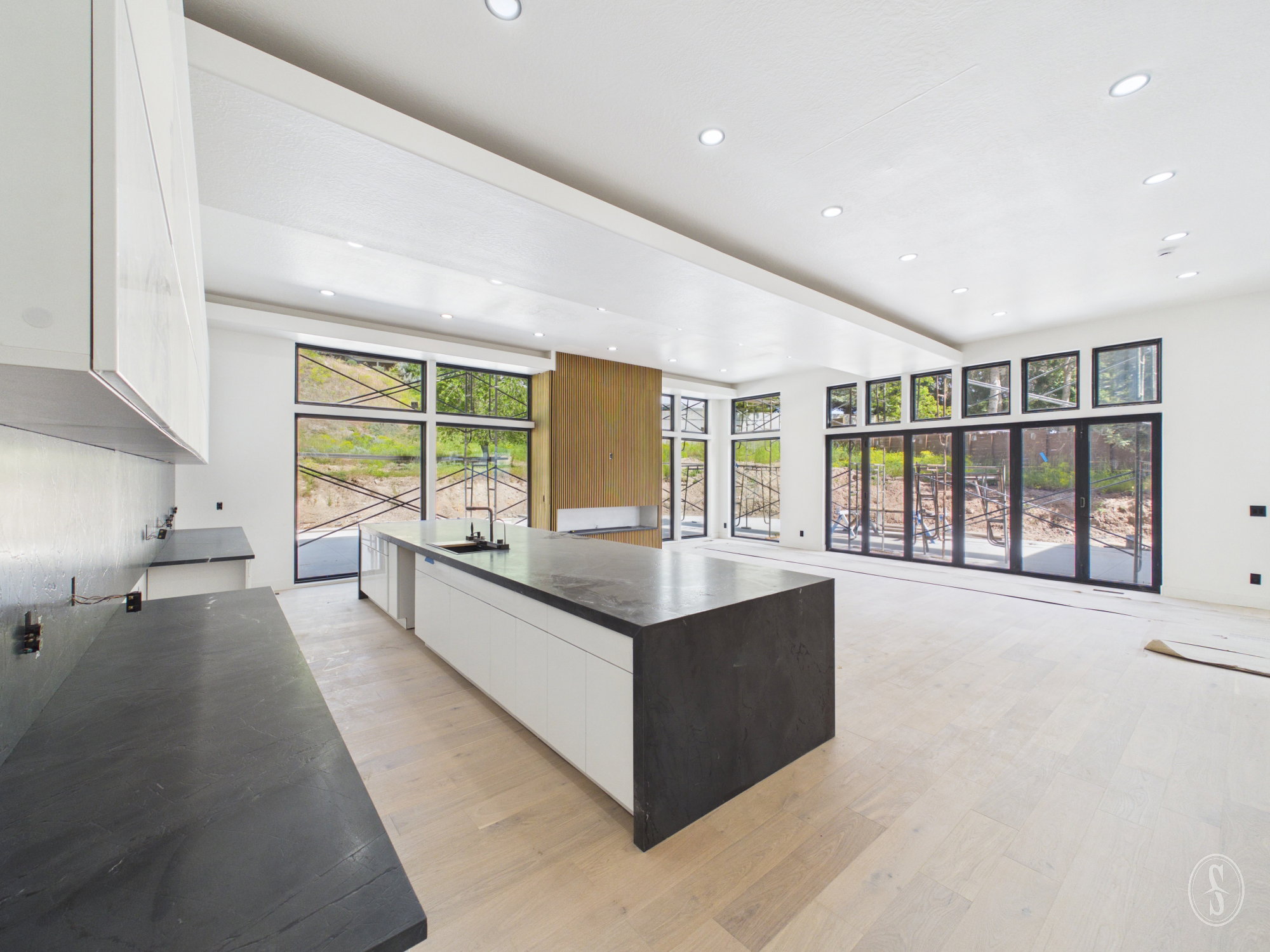 Spacious open-concept interior featuring light wood-finish flooring, a large kitchen island with a dark countertop, white cabinetry, and extensive black-framed windows across multiple walls