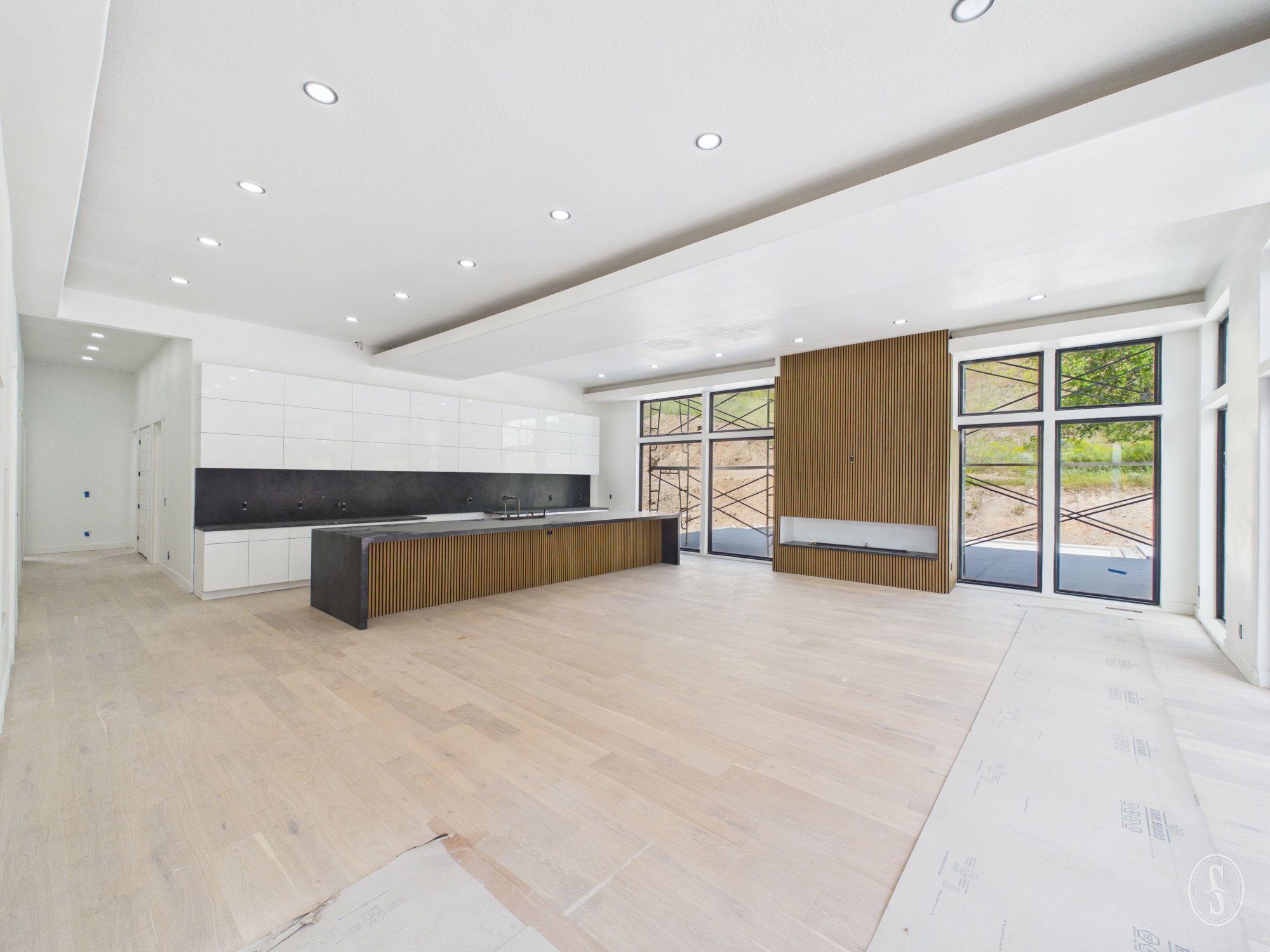 Expansive open-concept living space featuring light wood-finish flooring, a contemporary kitchen island with a dark countertop, white flat-panel cabinetry, and a full wall of floor-to-ceiling windows