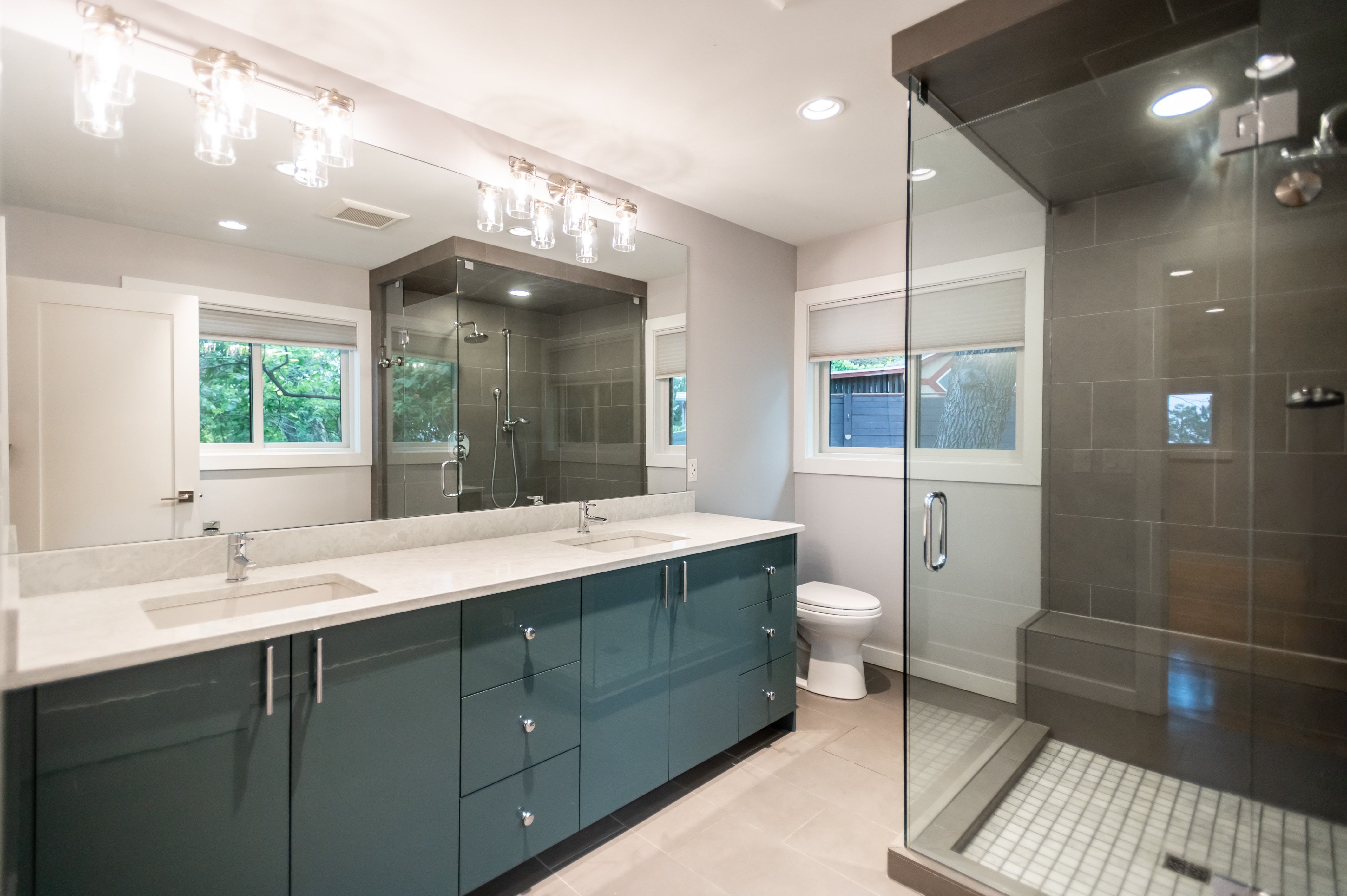 Bathroom featuring a dual vanity with a light-colored countertop and teal cabinetry, a large frameless mirror, a spacious glass-enclosed shower with a bench, and recessed lighting