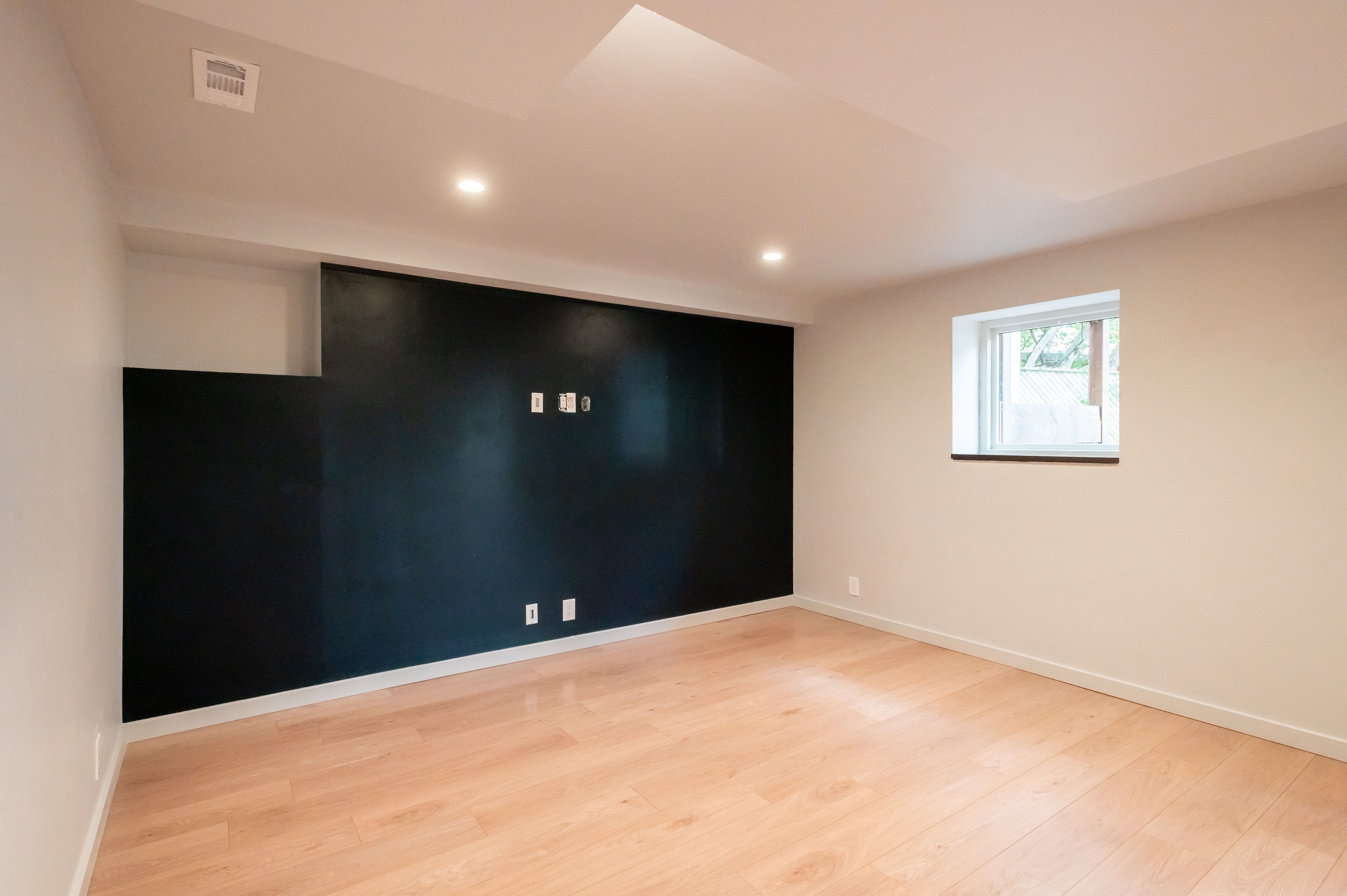 Bright room featuring light wood-finish flooring, a matte black accent wall with media outlets, recessed lighting, white baseboards, and a single window with white trim