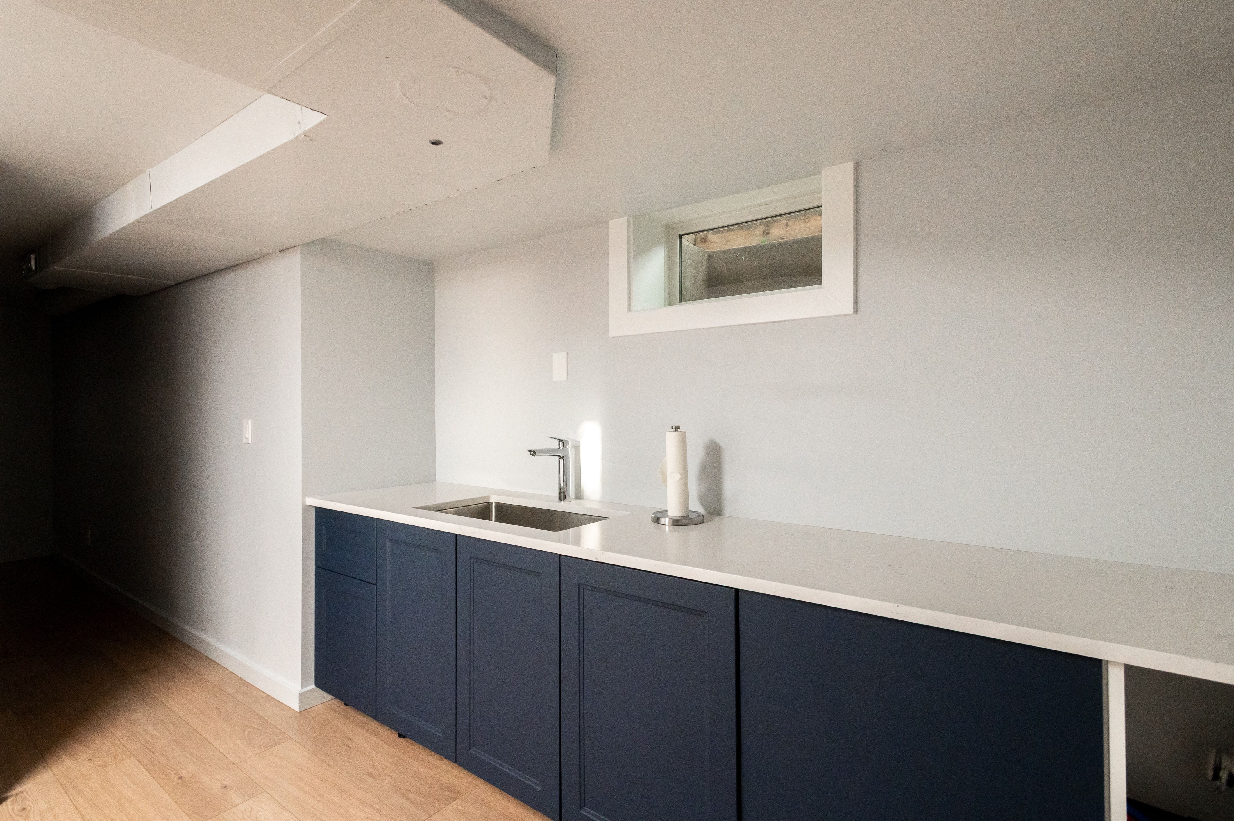 Compact kitchenette featuring a stainless steel sink, contemporary faucet, dark blue cabinetry, light-toned countertop, and light wood-finish flooring
