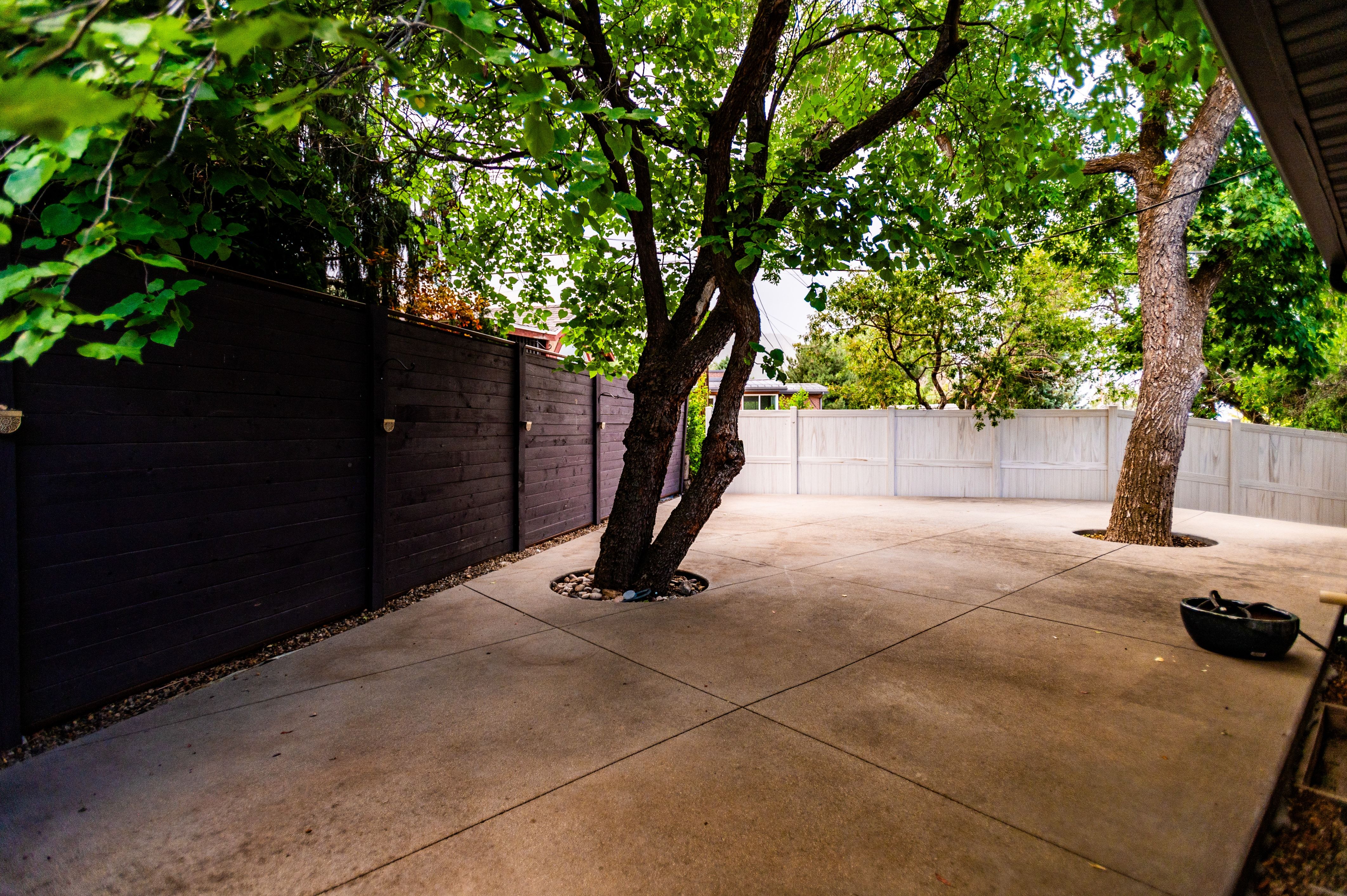Spacious concrete patio with mature trees, surrounded by a dark-stained wood privacy fence and a light-colored panel fence