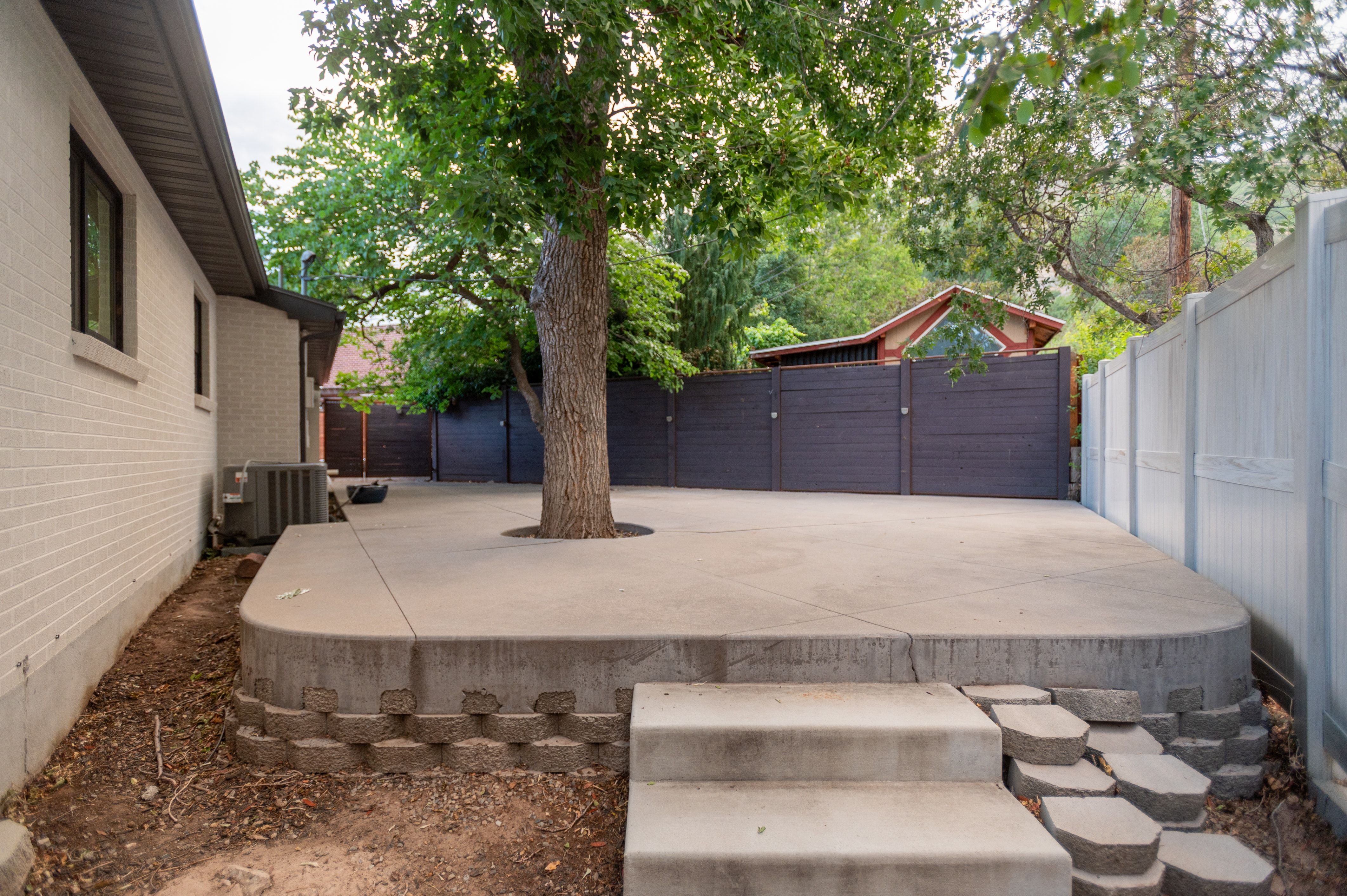 Expansive concrete patio with integrated mature tree, surrounded by a dark wood-finish privacy fence and a white vinyl privacy fence