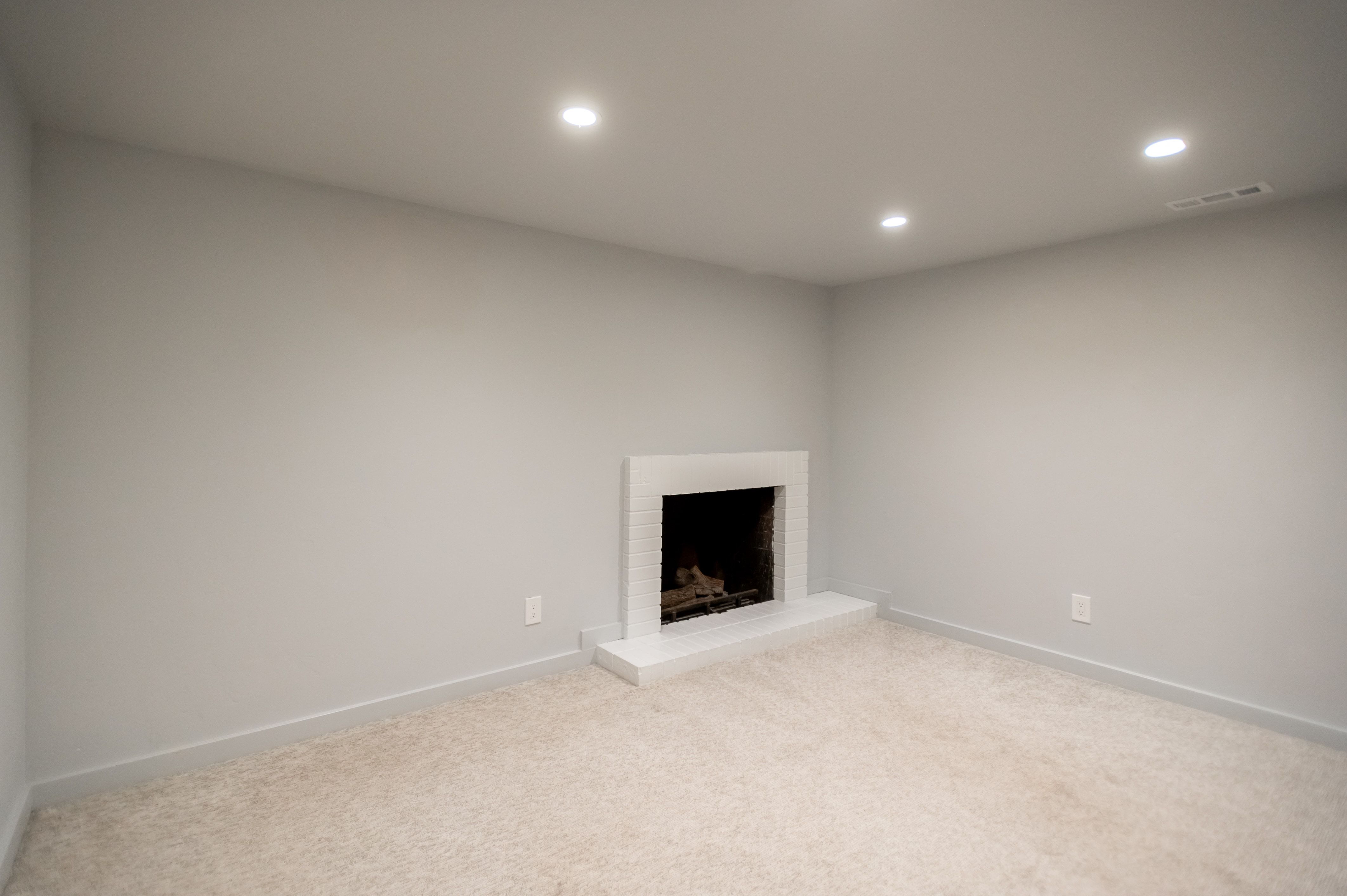 Recessed lighting illuminates a space featuring a white brick fireplace with a hearth, light neutral-toned carpeting, and light gray walls