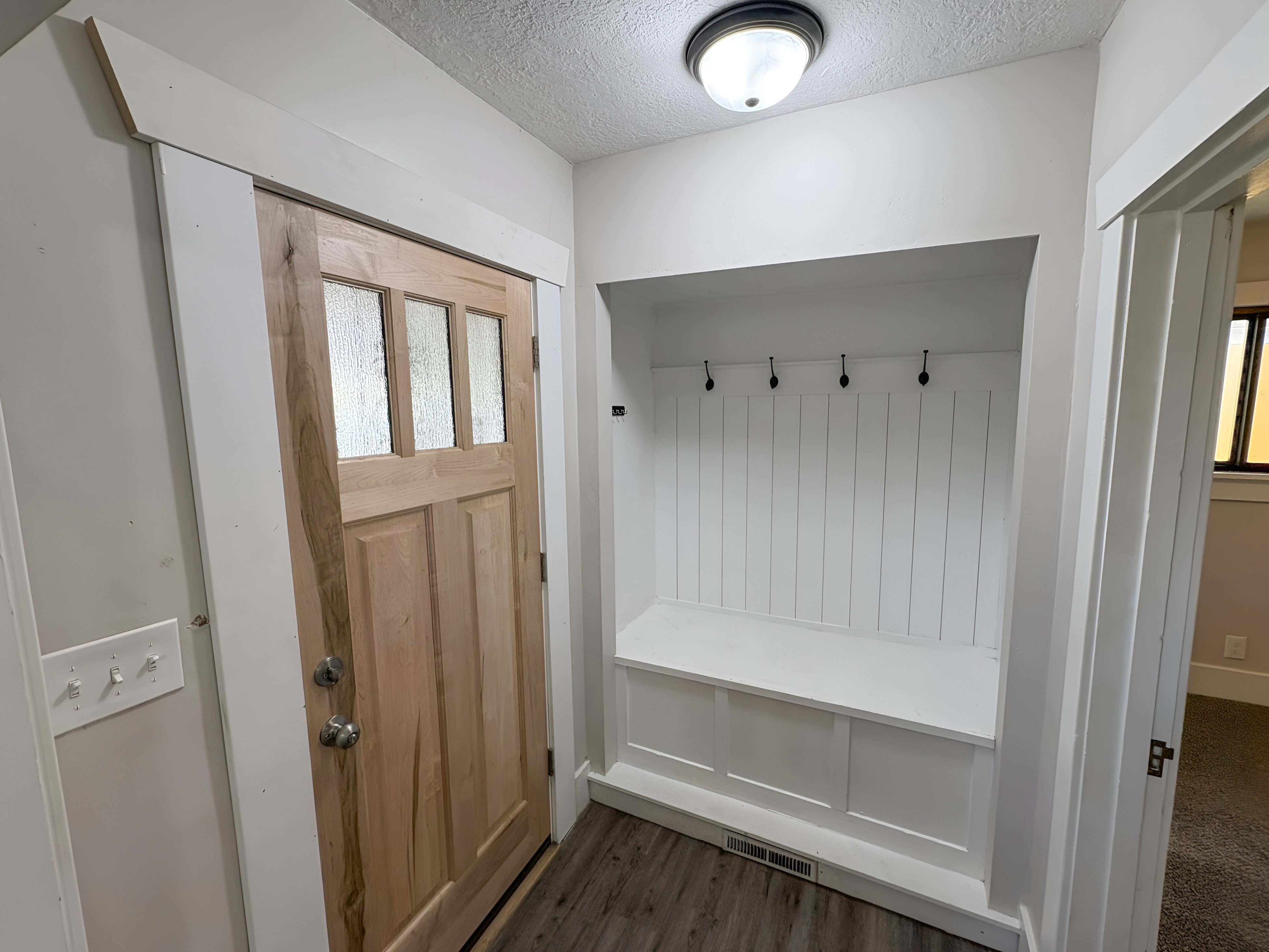 Entryway featuring a natural wood door with frosted glass panels, wood-finish flooring, and a built-in white mudroom bench with beadboard backing and black coat hooks
