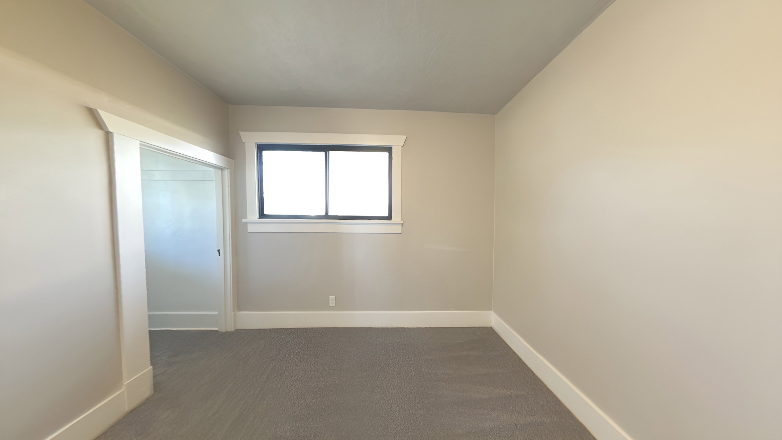 Neutral-toned room featuring a single window with dark framing and white trim, complemented by light gray carpeting and white baseboards
