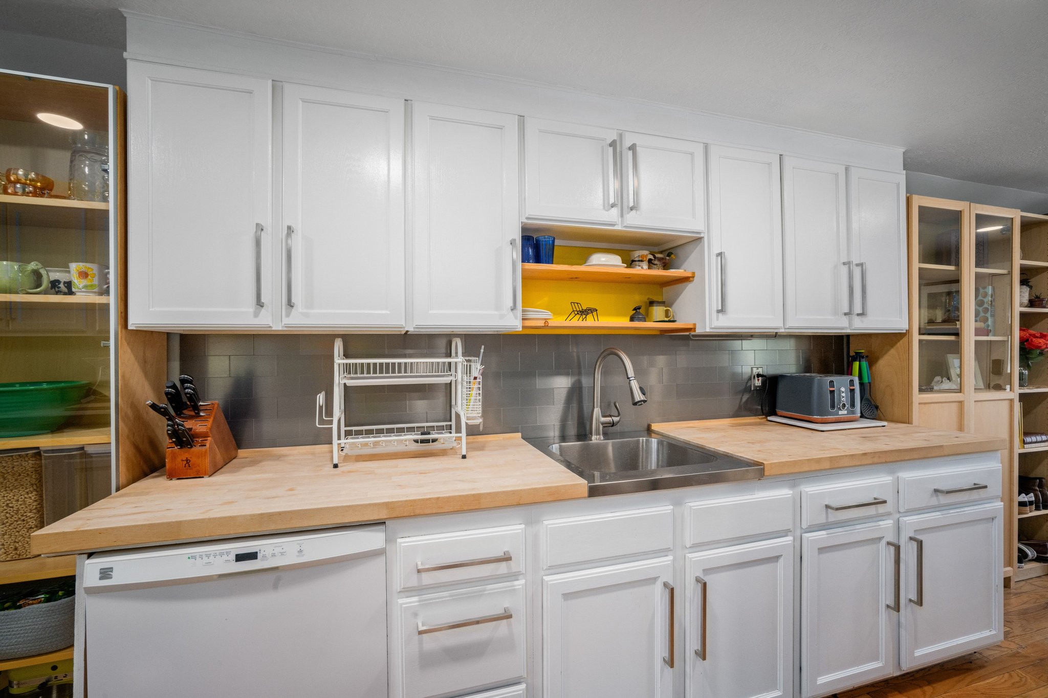 Contemporary kitchen featuring white cabinetry with brushed nickel hardware, wood-finish countertops, stainless steel sink with a gooseneck faucet, a tile backsplash, and a built-in dishwasher
