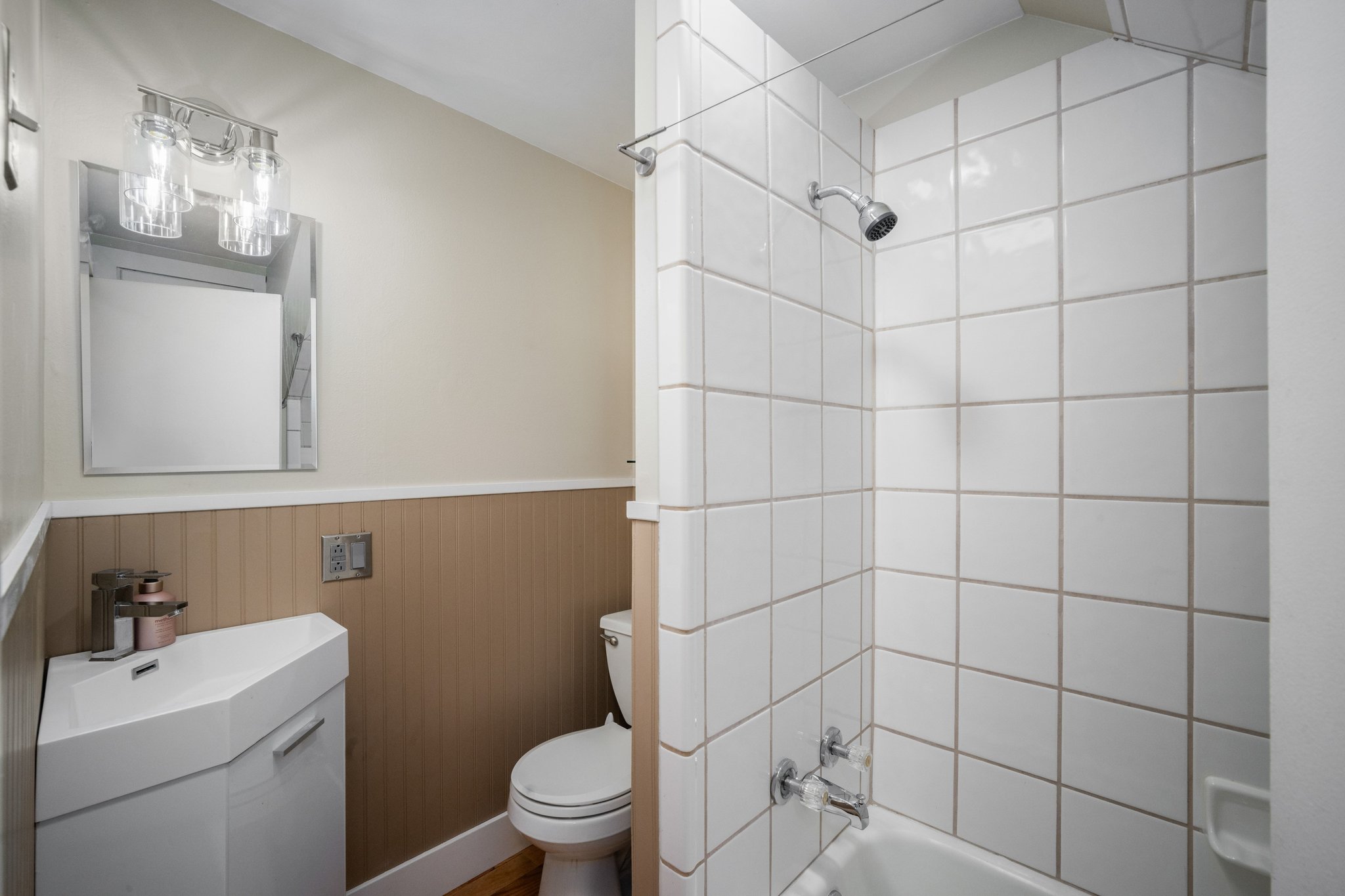 Bathroom featuring a white tile shower surround, a corner vanity with integrated sink, beadboard wainscoting, and wood-finish flooring