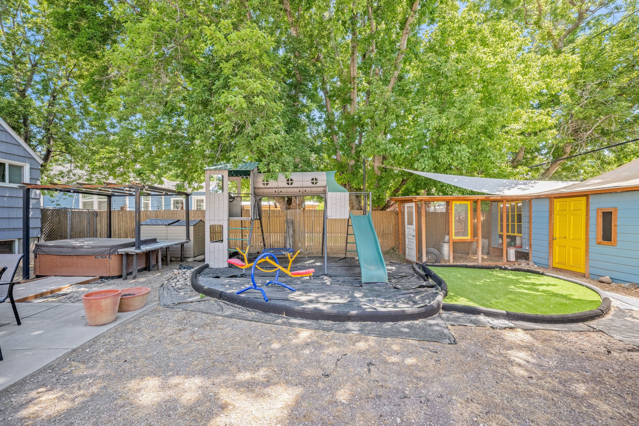 Expansive backyard featuring a built-in hot tub under a pergola, a play structure with a slide and swings, a dedicated artificial turf area, and a blue-sided outbuilding with a yellow door