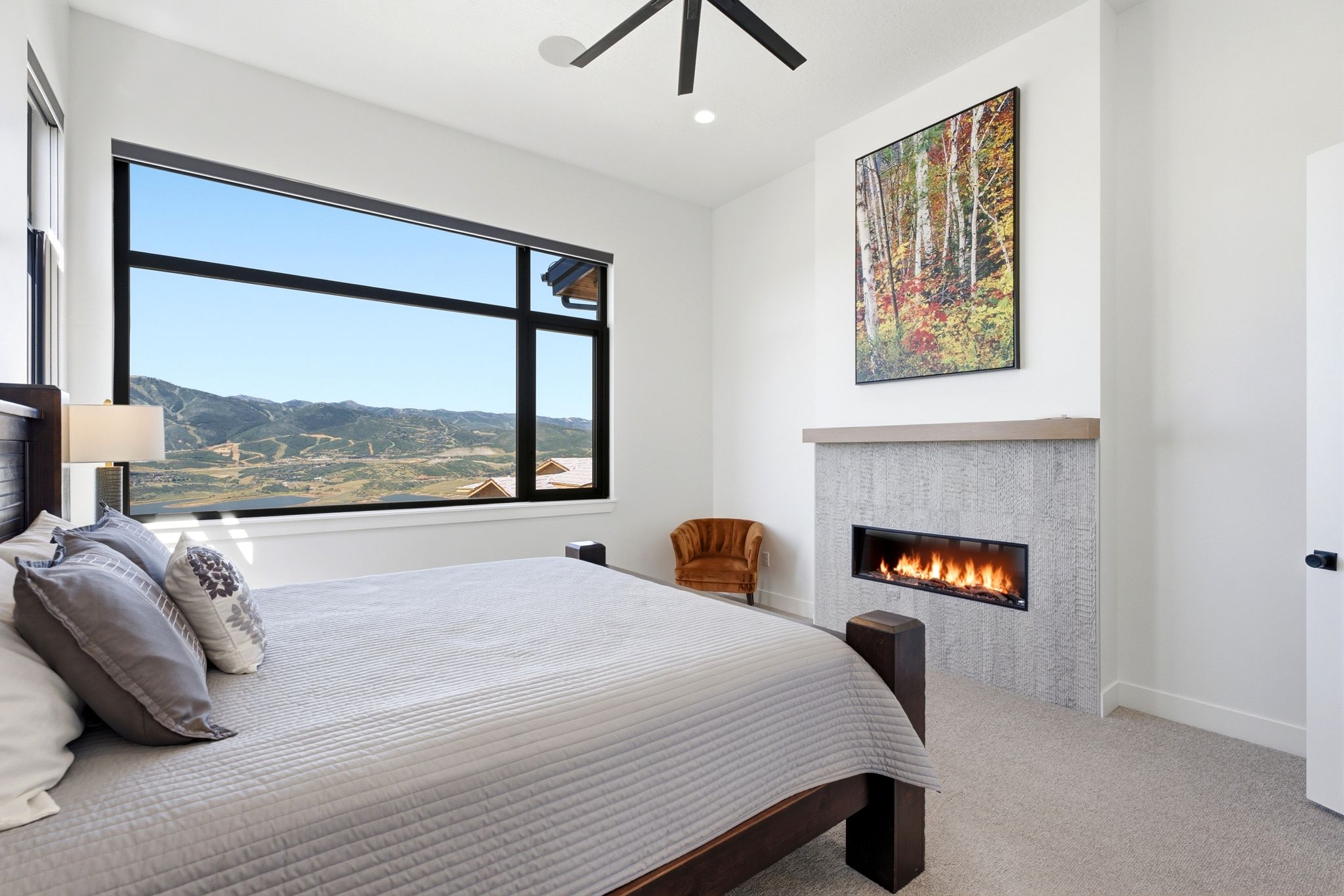 Spacious bedroom featuring an expansive horizontal window with mountain scenery, a linear gas fireplace with a textured tile surround, a wood mantle, and light-toned carpeting