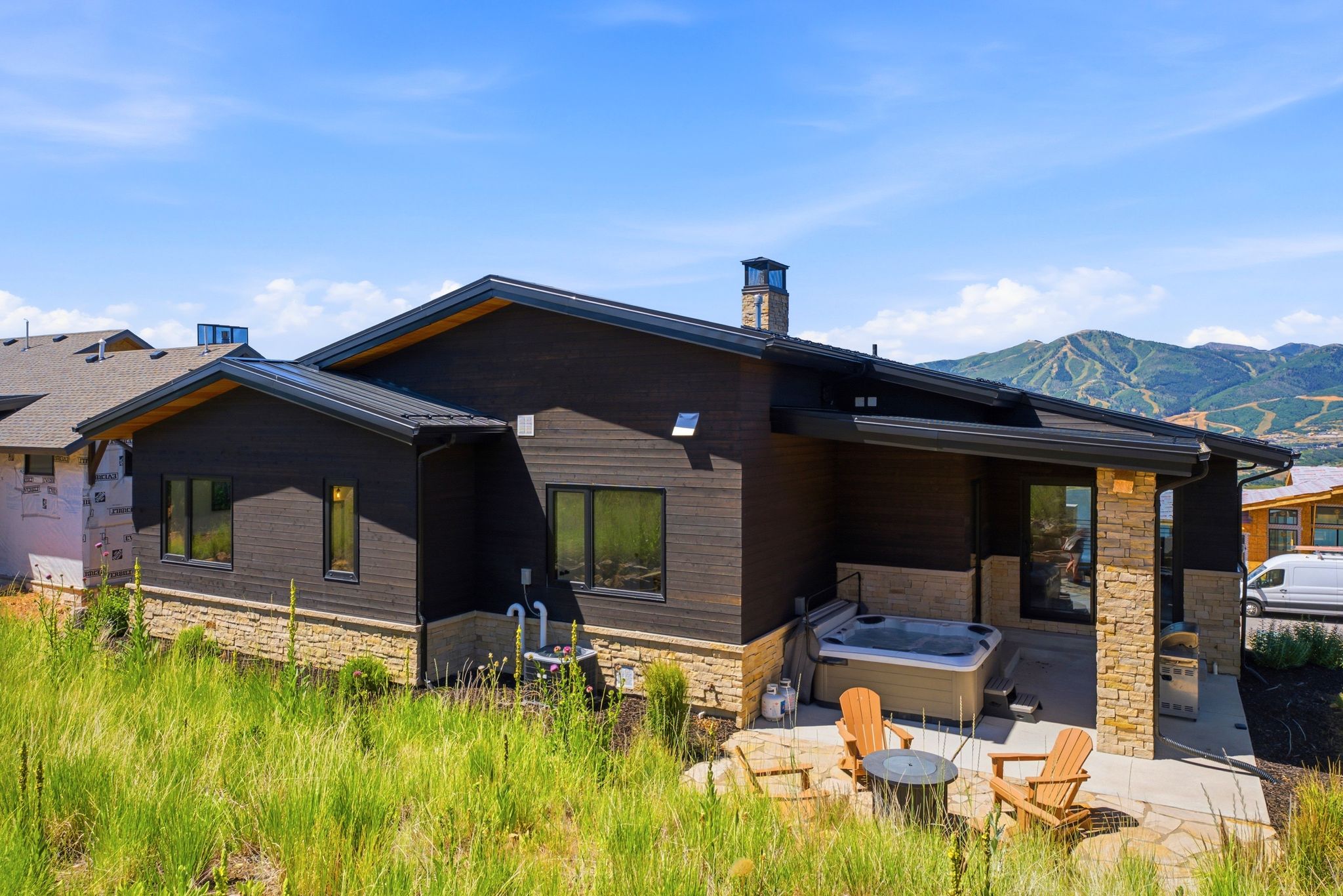 Contemporary rear exterior featuring dark wood-finish siding, stone accents, a metal roof, an outdoor patio with a built-in hot tub, and mountain scenery