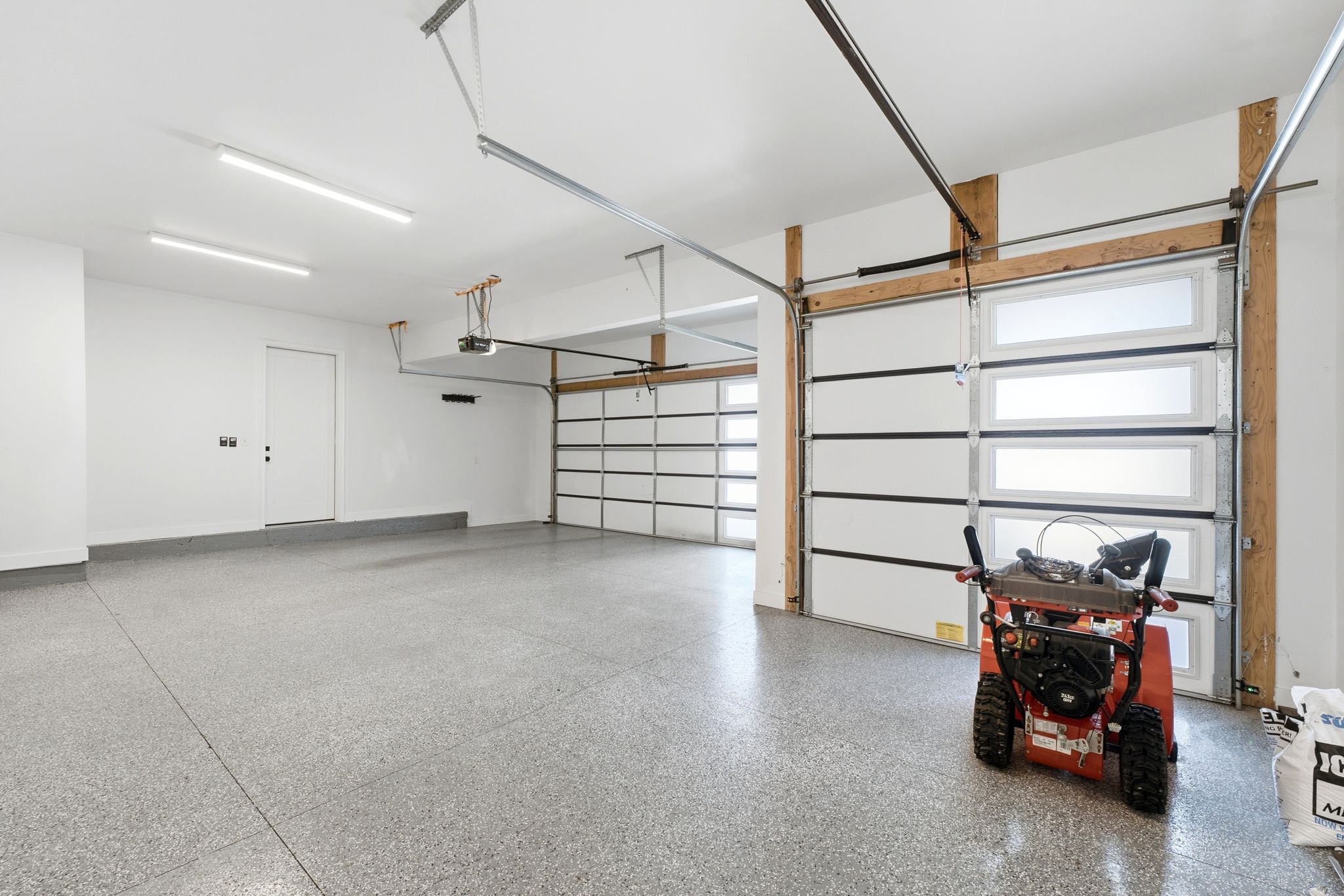 Spacious garage featuring a durable epoxy floor, white walls, two contemporary garage doors with frosted glass panels, overhead lighting, and an interior access door