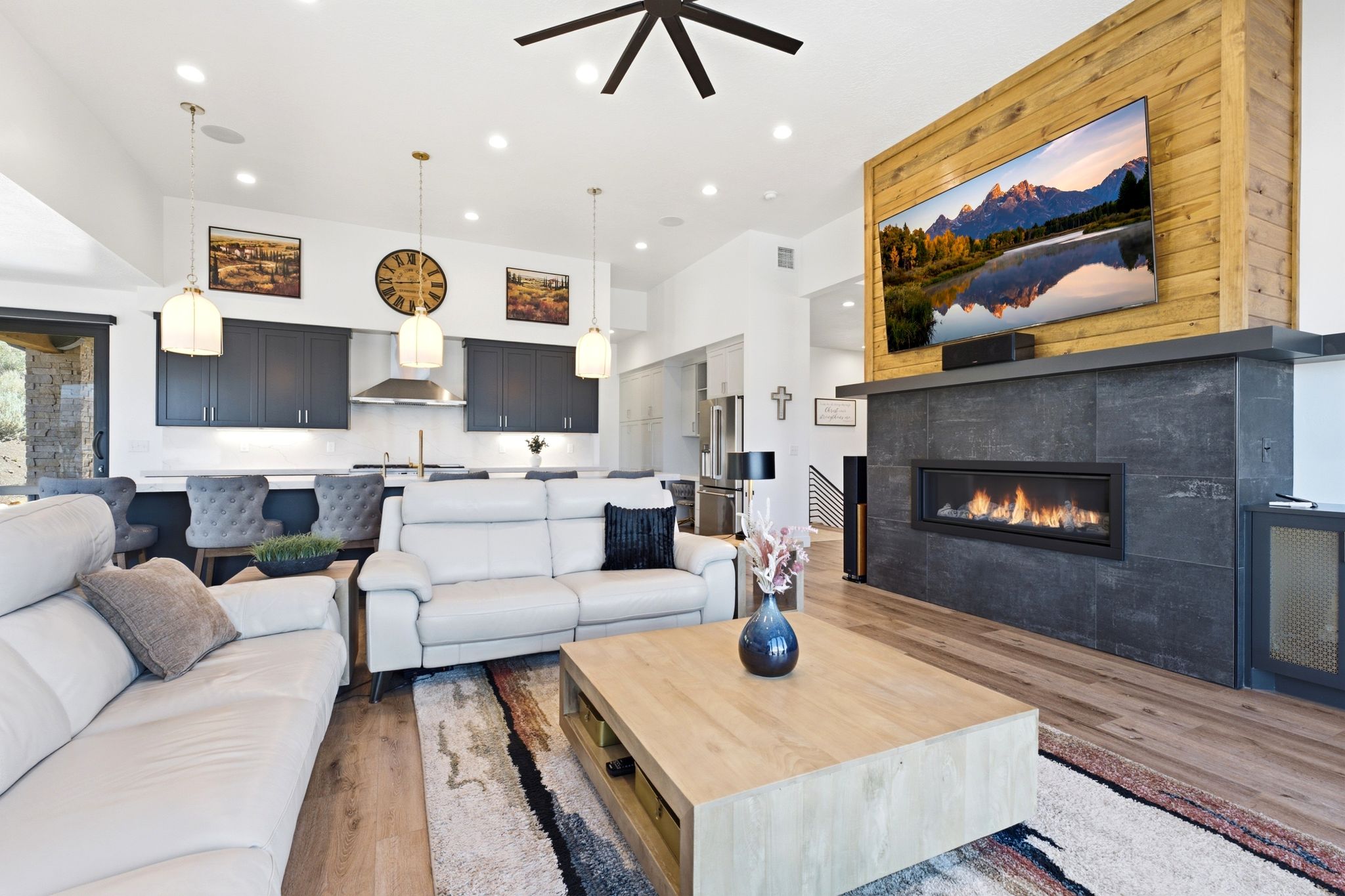 Open-concept interior featuring wood-finish flooring, a linear fireplace with a stone surround, and a kitchen with dark cabinetry and pendant lighting