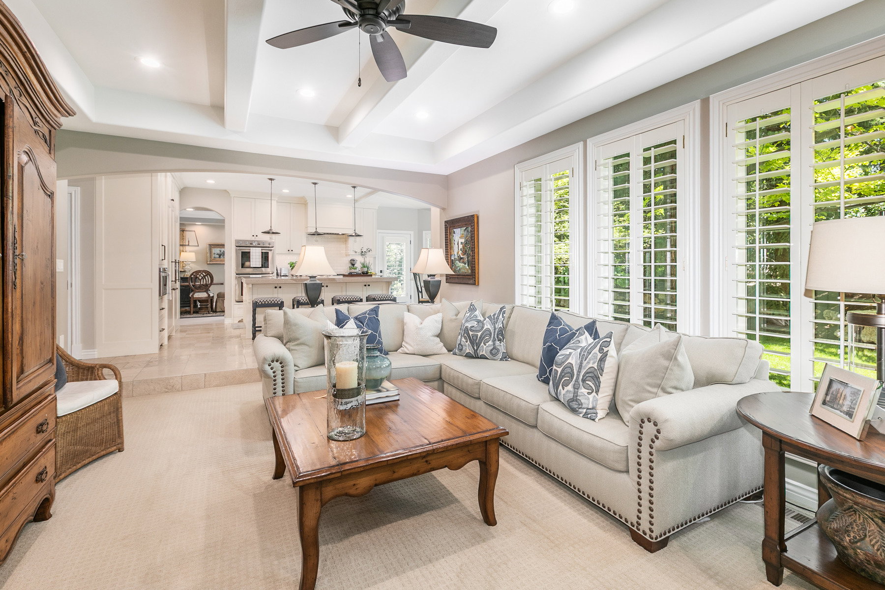 Spacious living area featuring coffered ceilings with recessed lighting, an arched entry to a kitchen with pendant lighting, and plantation shutters