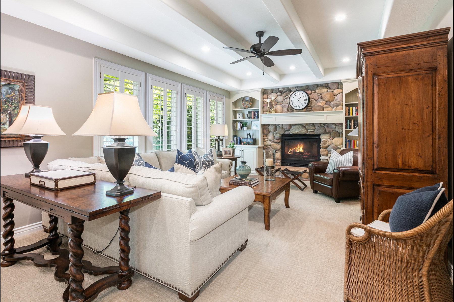 Spacious living area featuring a stone-surround fireplace with mantel, built-in shelving, vaulted ceiling with exposed beams, recessed lighting, and plantation shutters
