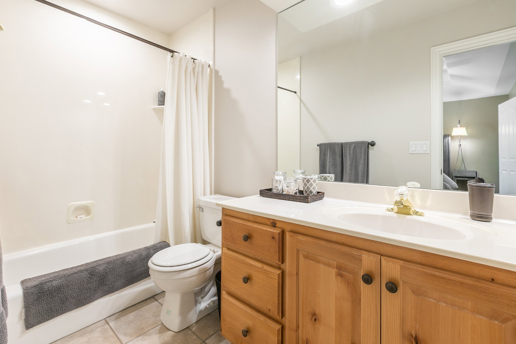 Bathroom featuring a light wood vanity with an integrated sink, a large wall-mounted mirror, a tub/shower combination with a shower curtain, a commode, and light-colored floor tiling