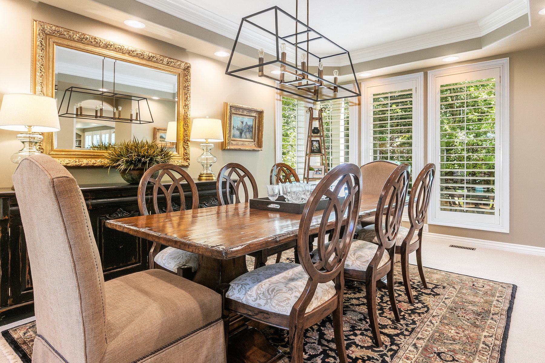 Dining area featuring a rectangular wood dining table, two large windows with white louvered shutters, a black metal chandelier, a decorative area rug, and tray ceiling detail