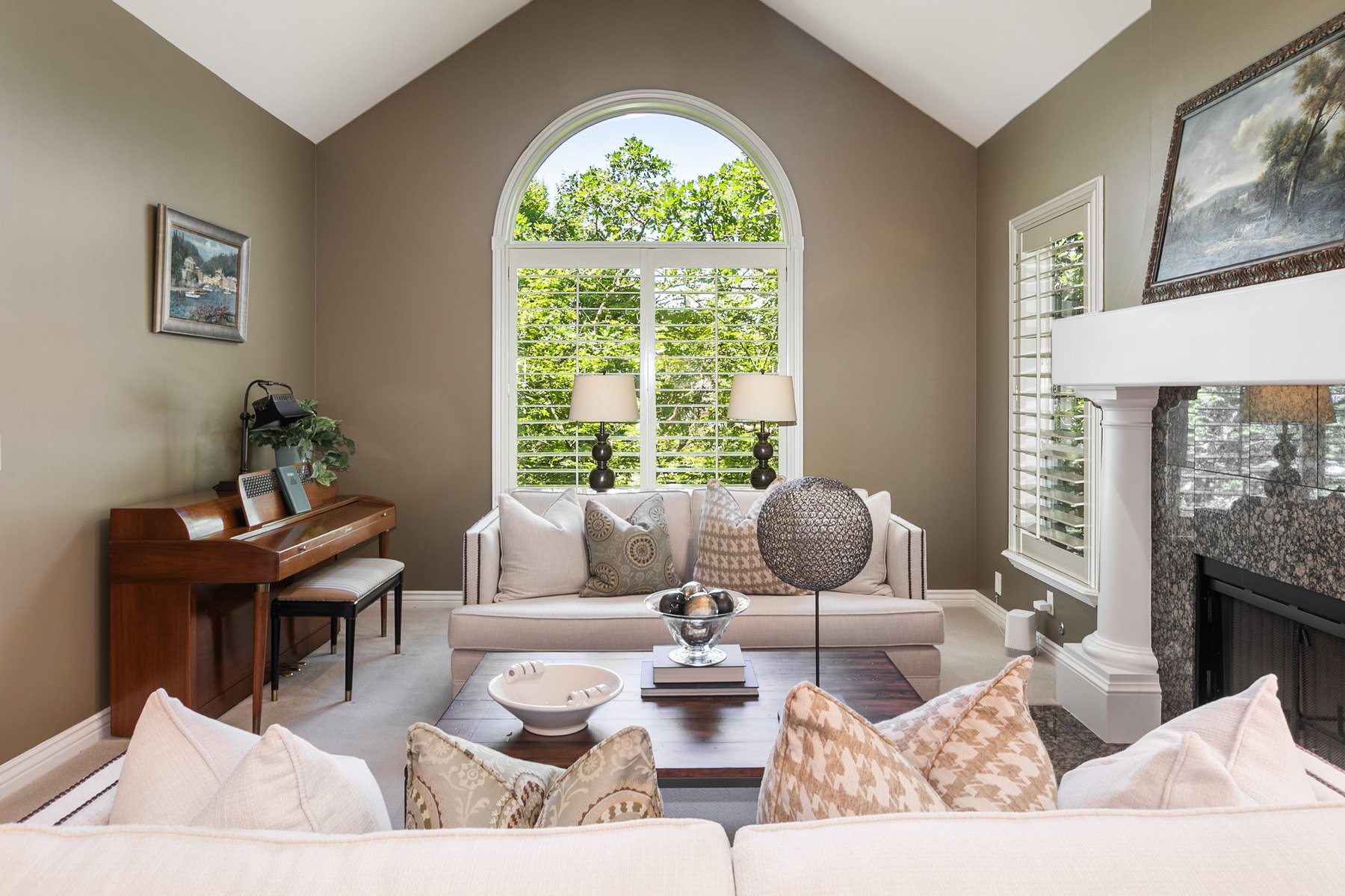 Vaulted ceiling with an arched window, plantation shutters, and a fireplace featuring a stone surround and a substantial mantel