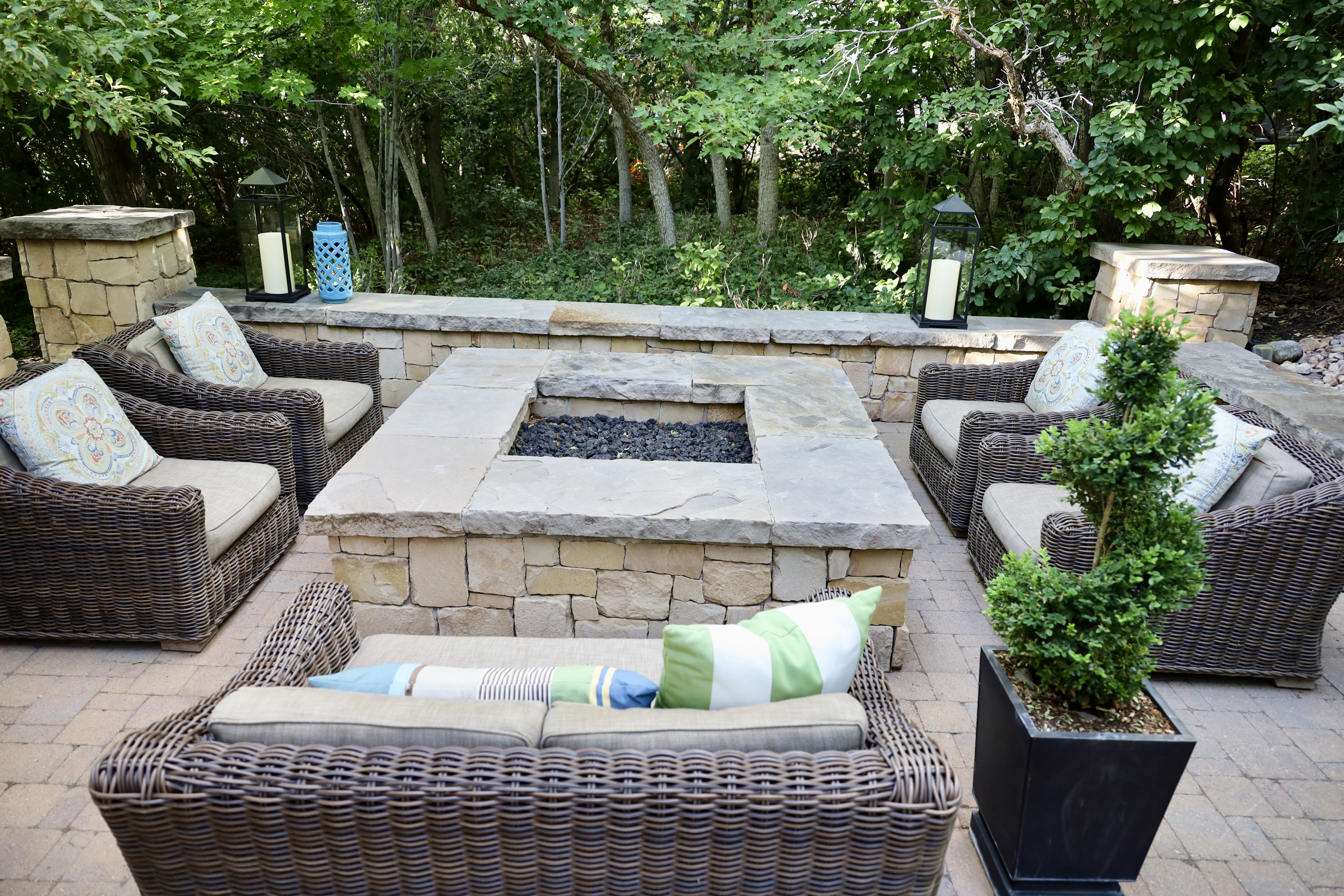 Paver patio featuring a stone-clad fire pit with a stone cap, surrounded by a stone retaining wall and lush greenery