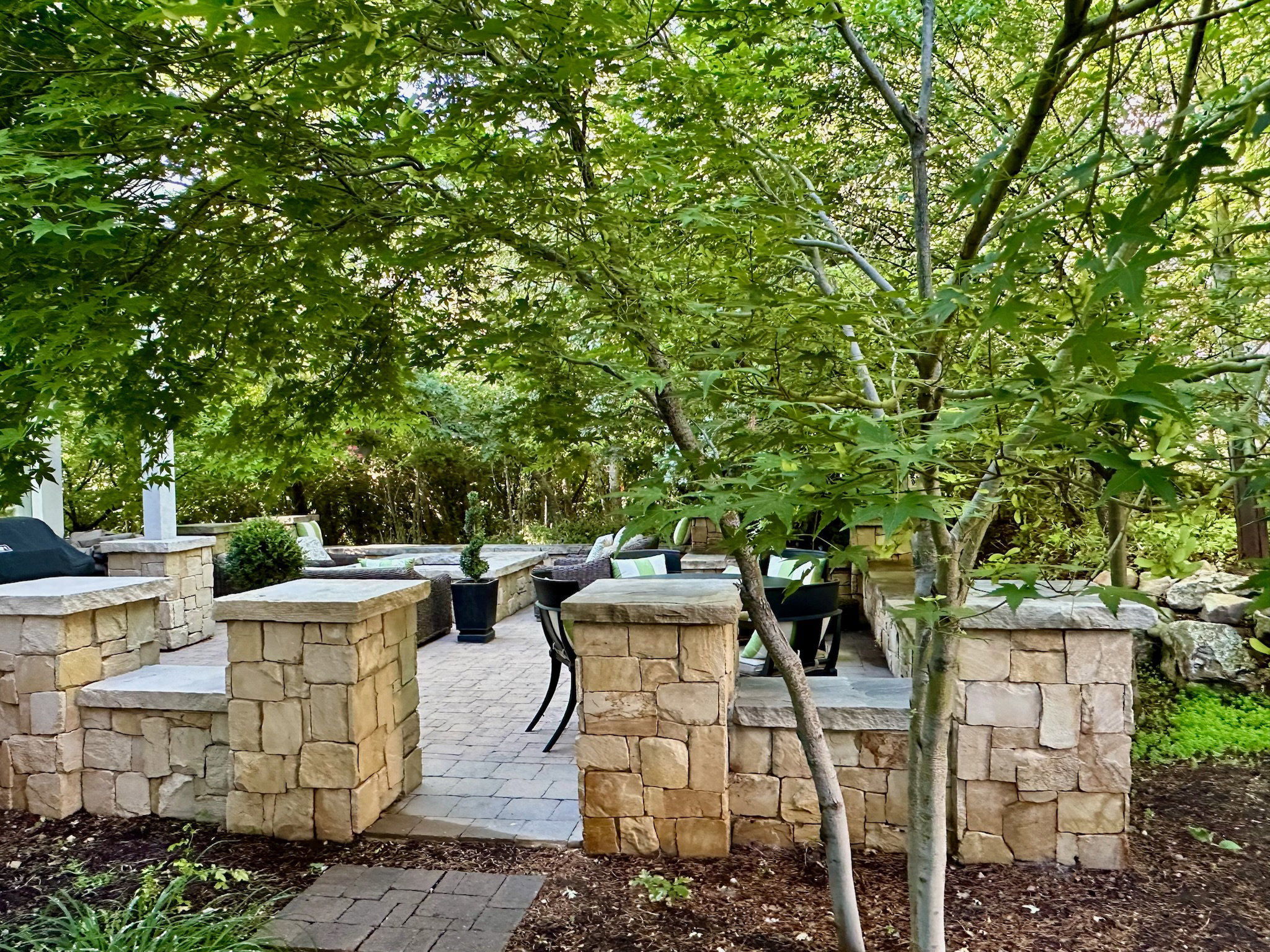 Outdoor patio featuring extensive stone wall hardscaping, paver flooring, mature trees providing natural shade, and built-in seating ledges