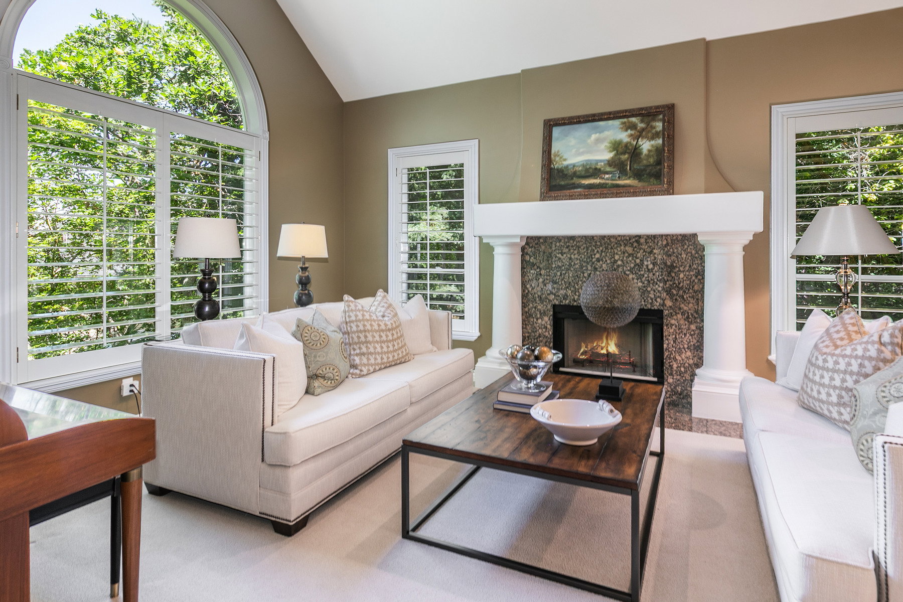 Living area featuring a stone-faced fireplace with a substantial mantel, flanked by a large arched window and additional windows with plantation shutters