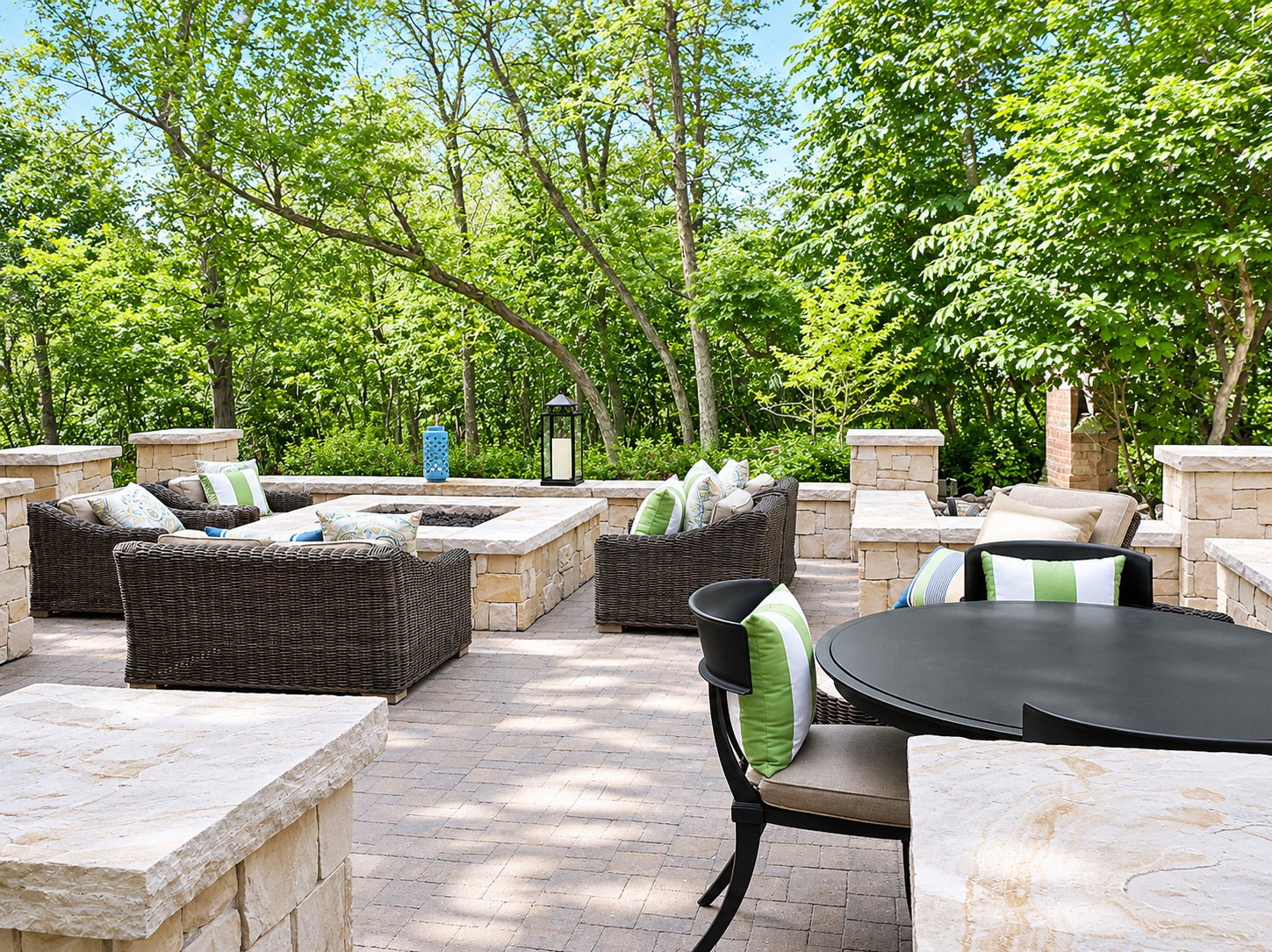 Expansive paver patio featuring a built-in fire pit with stone surround, perimeter stone walls, and mature tree landscaping