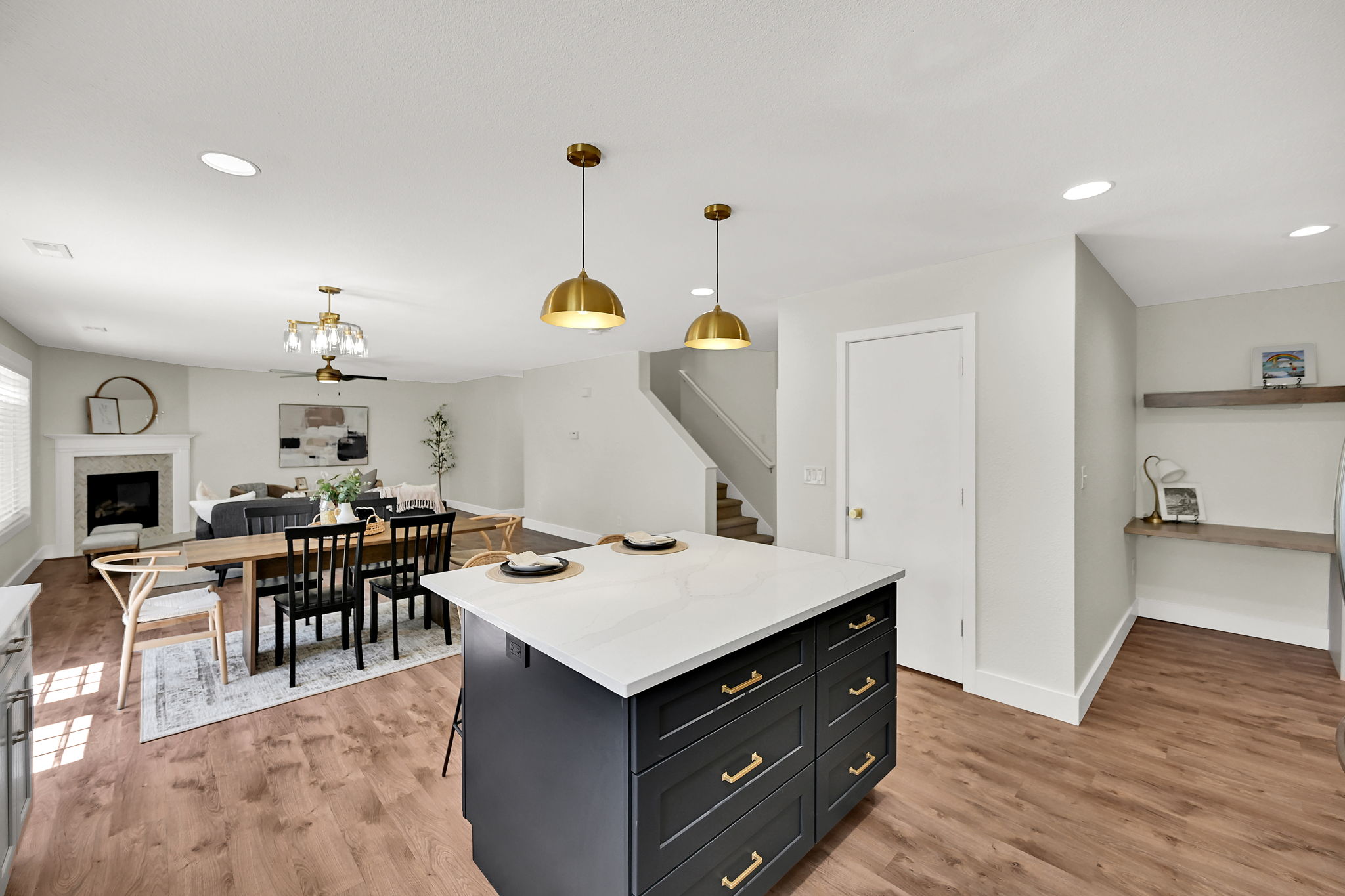 Spacious open-concept living area featuring a kitchen island with a light-toned countertop, gold pendant lighting, wood-finish flooring, a fireplace with a substantial mantel, and built-in floating shelves