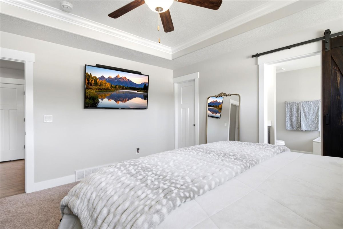 Recessed ceiling with crown molding, ceiling fan with light fixture, light gray wall color, barn door entry, and carpeted flooring