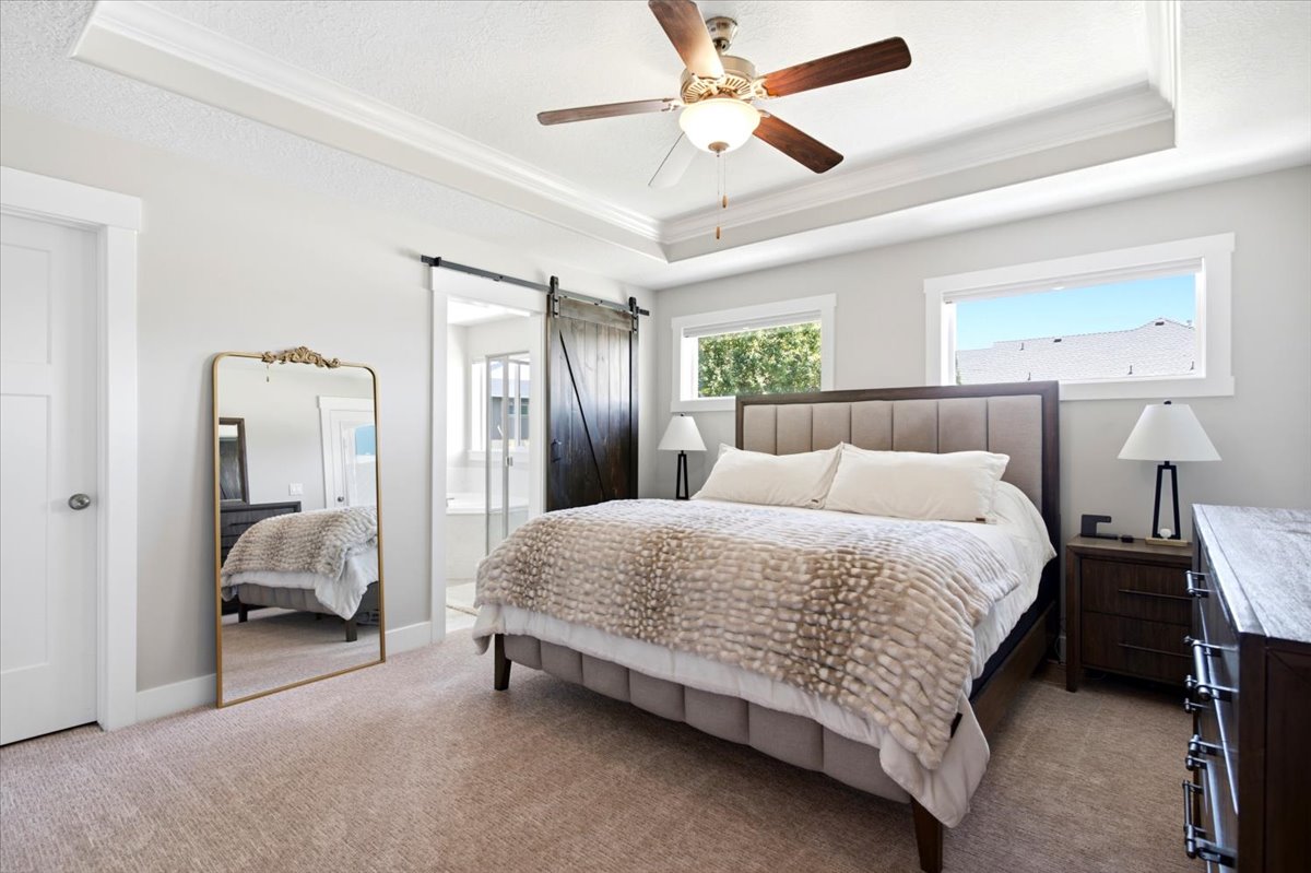 Spacious bedroom featuring a tray ceiling with a ceiling fan, two windows, and light-colored carpeting