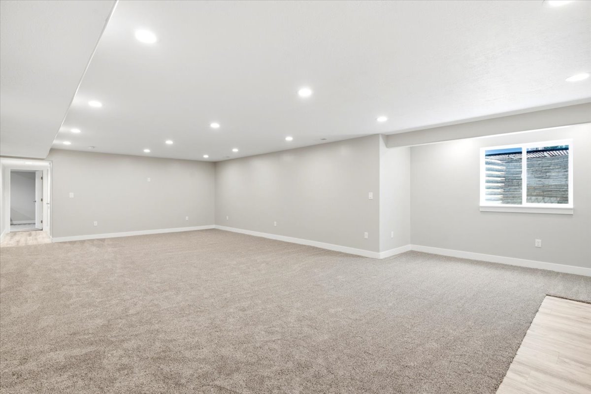 Expansive carpeted room featuring recessed lighting, a window with a stone-look exterior, light gray walls, white baseboards, and a doorway leading to a space with wood-finish flooring