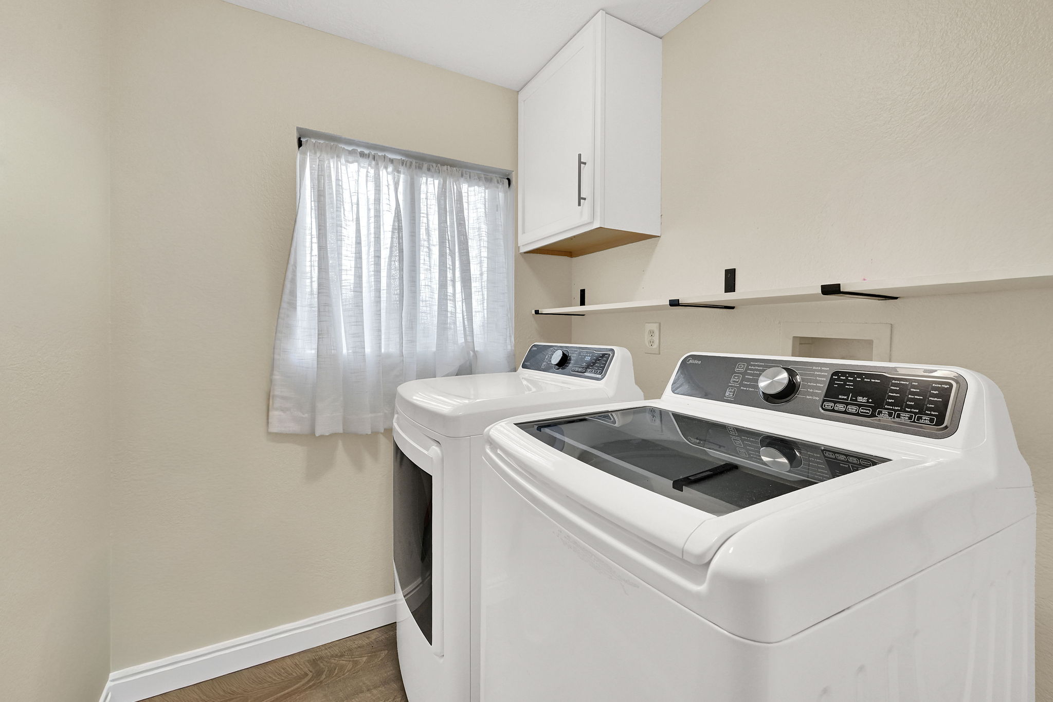 Main floor laundry area  off the kitchen featuring wood-finish flooring, a window with curtains, a built-in wall cabinet, and open shelving (Washer and Dryer stay with the home)