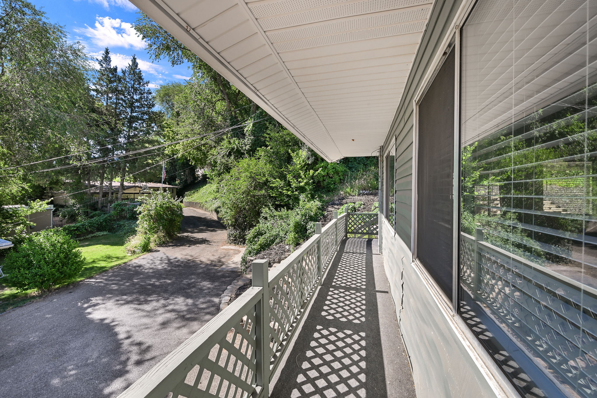 Covered porch with lattice railing overlooking a paved driveway, surrounded by mature trees and natural landscaping