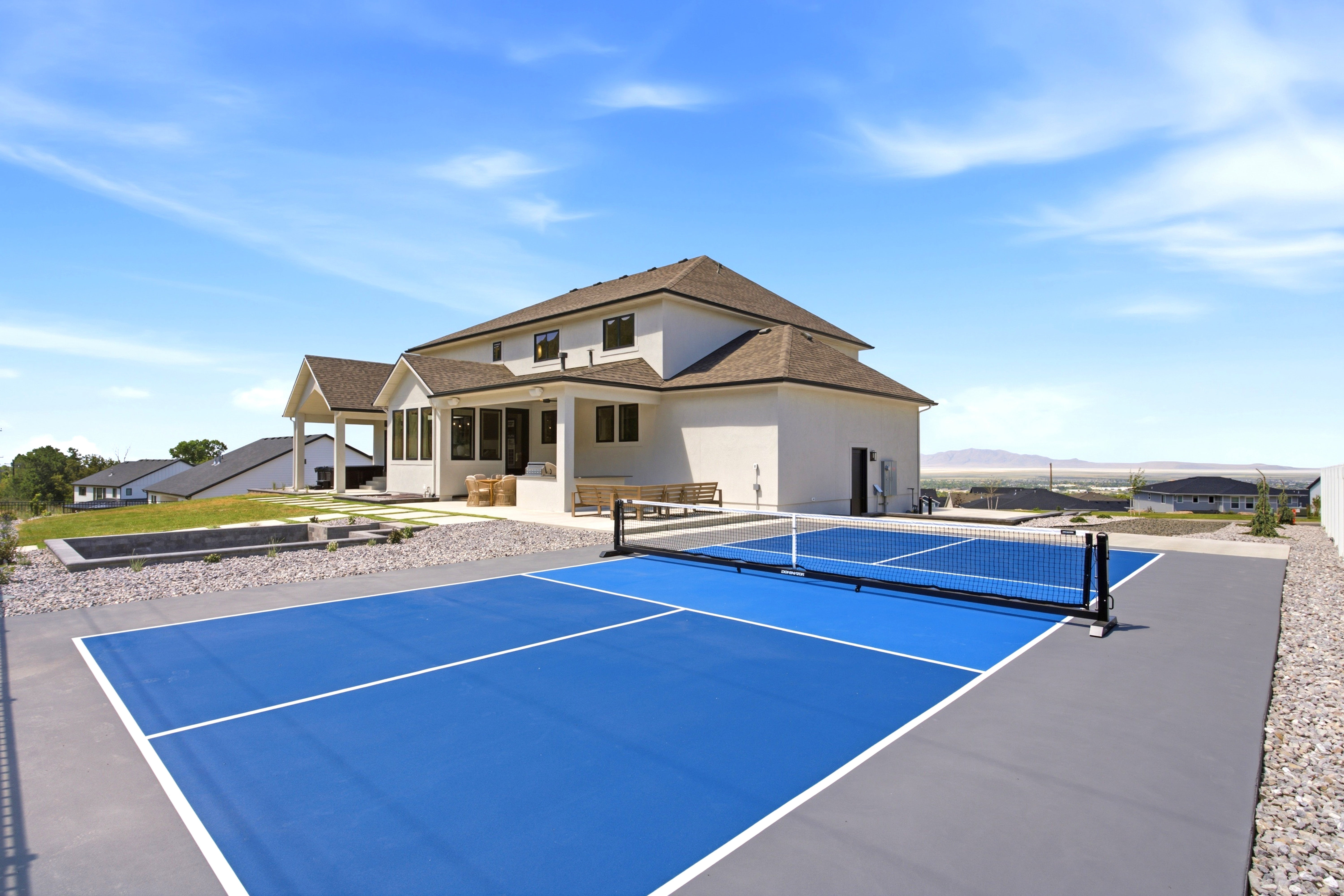 Expansive rear exterior featuring a dedicated blue-surfaced pickleball court, a stone-lined planter with greenery, and a covered patio area