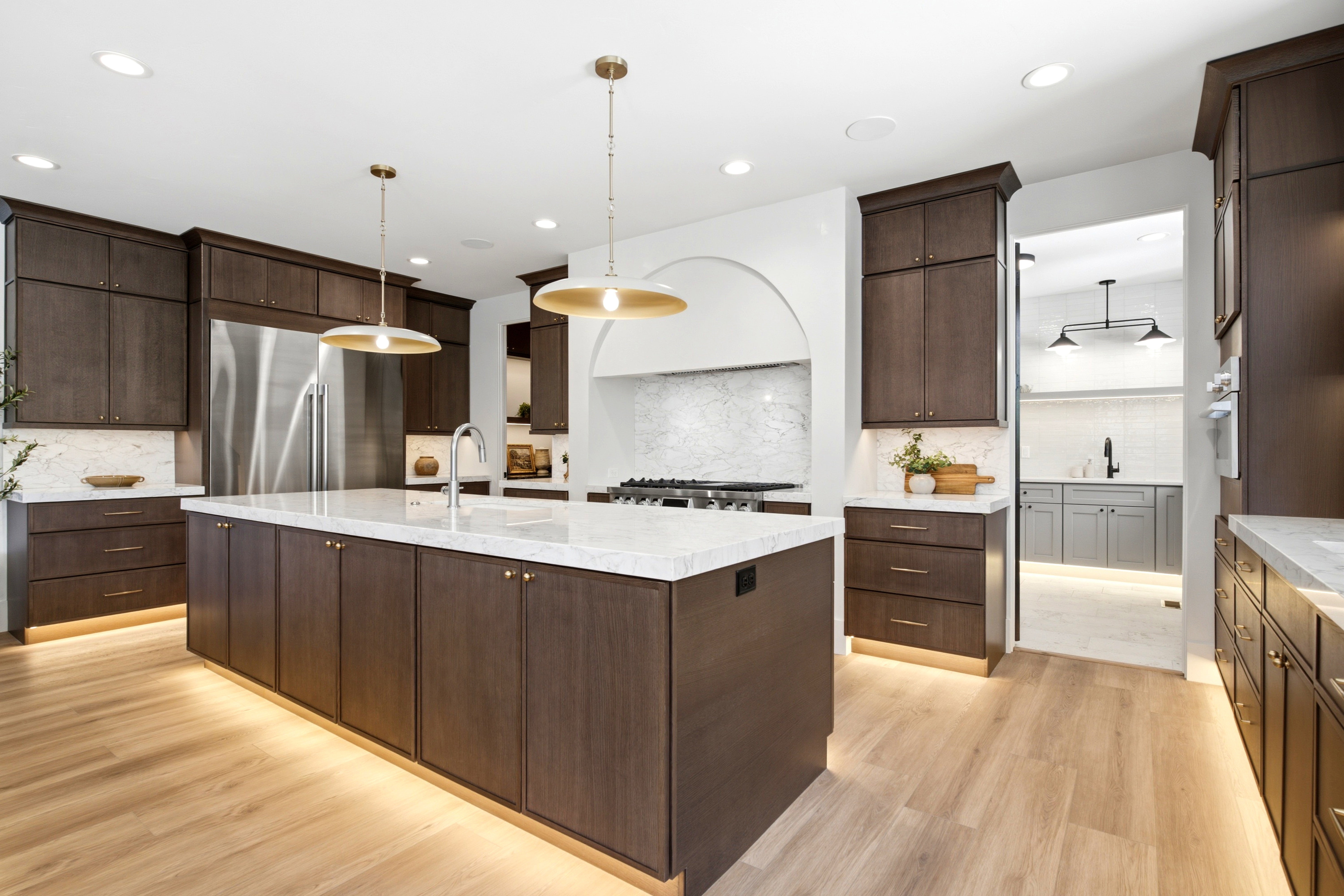 Gourmet kitchen featuring a large central island with integrated sink, light wood-finish flooring, dark wood cabinetry with under-cabinet lighting, stainless steel appliances, and elegant pendant lighting