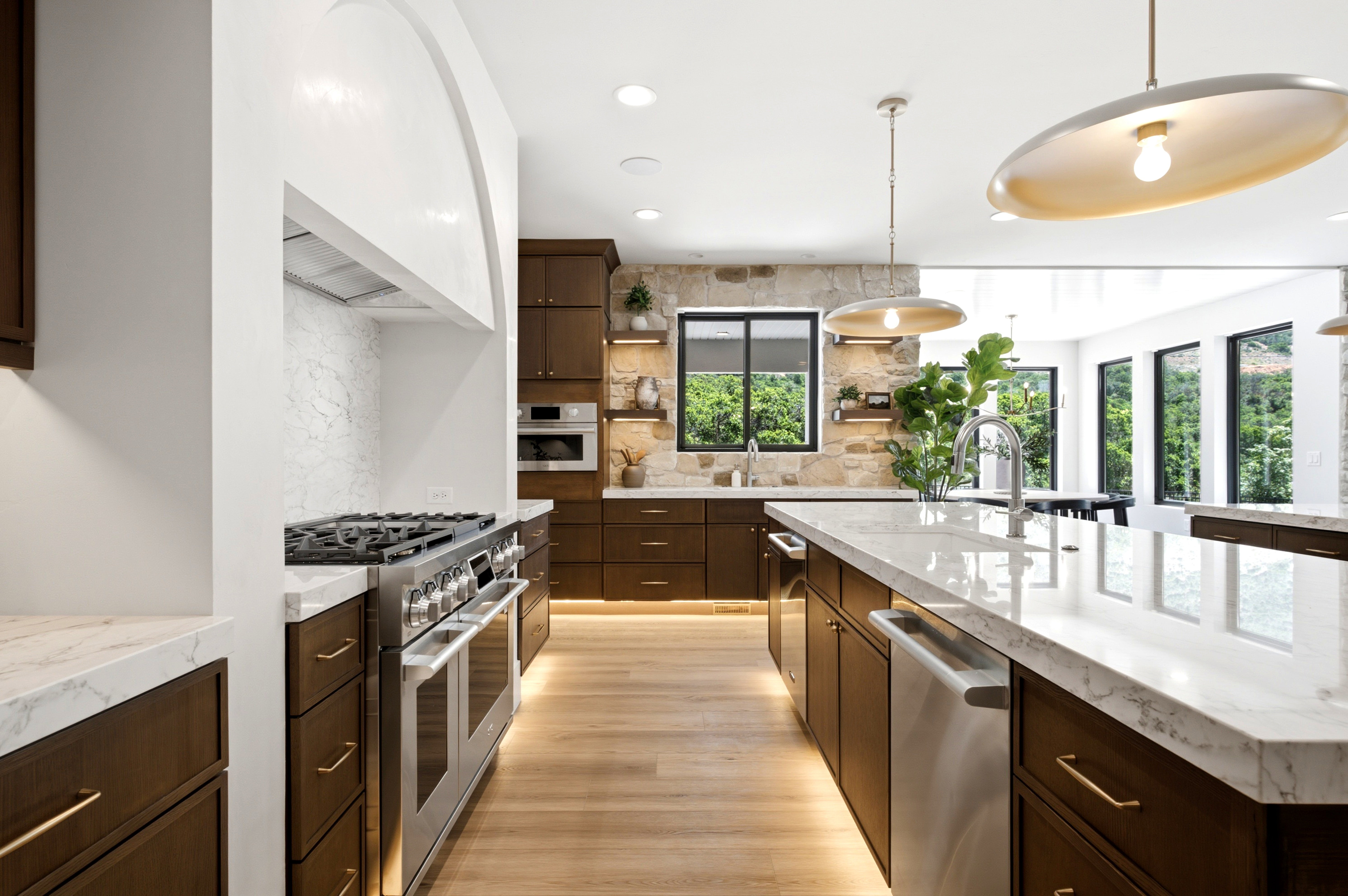 Gourmet kitchen featuring a professional-grade gas range, stone accent wall, large center island with integrated sink, wood-finish flooring, and ample cabinetry with under-cabinet lighting