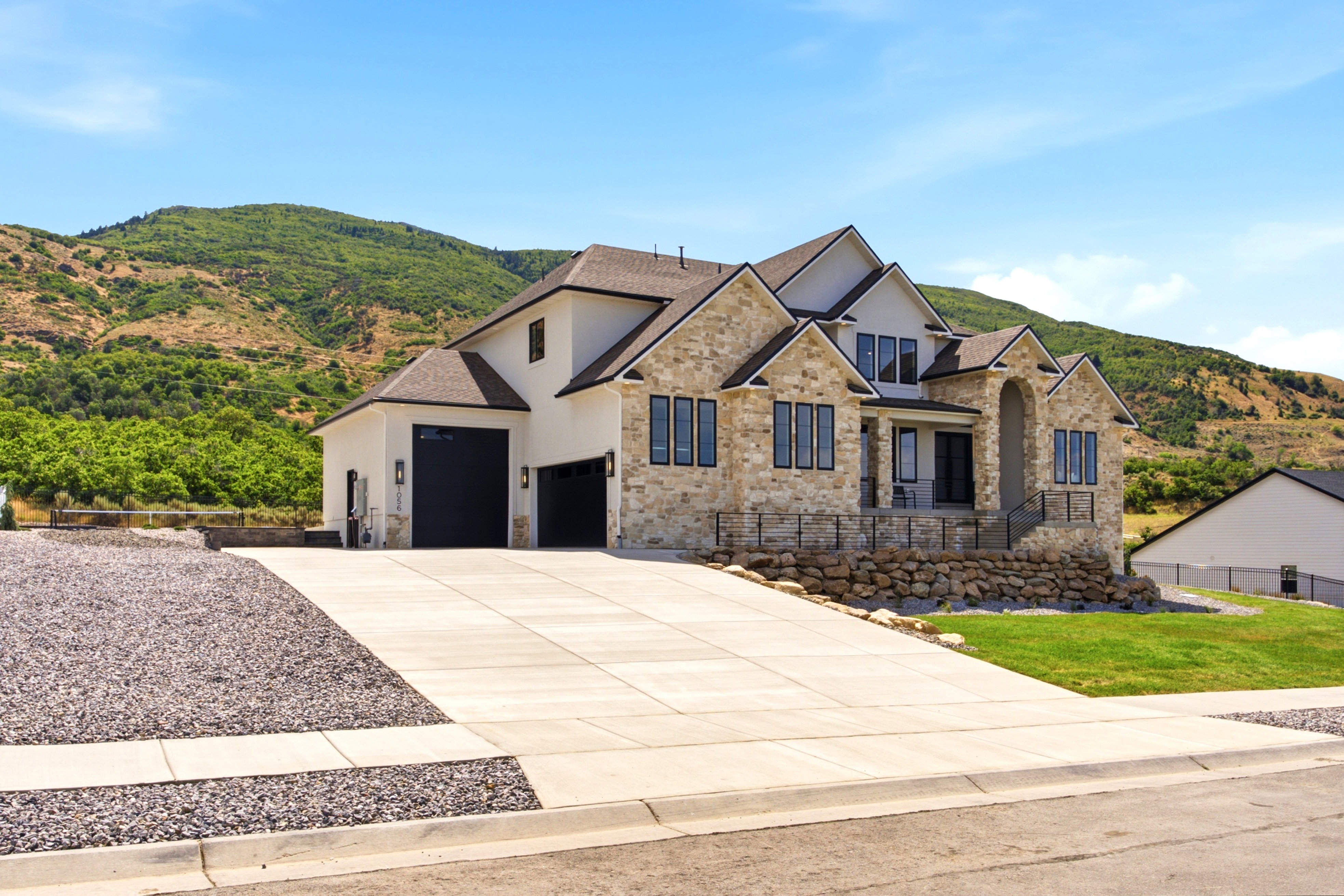 Contemporary architectural design featuring stone and stucco exterior, prominent gables, a large concrete driveway, two dark garage doors, and a front porch with a black metal railing