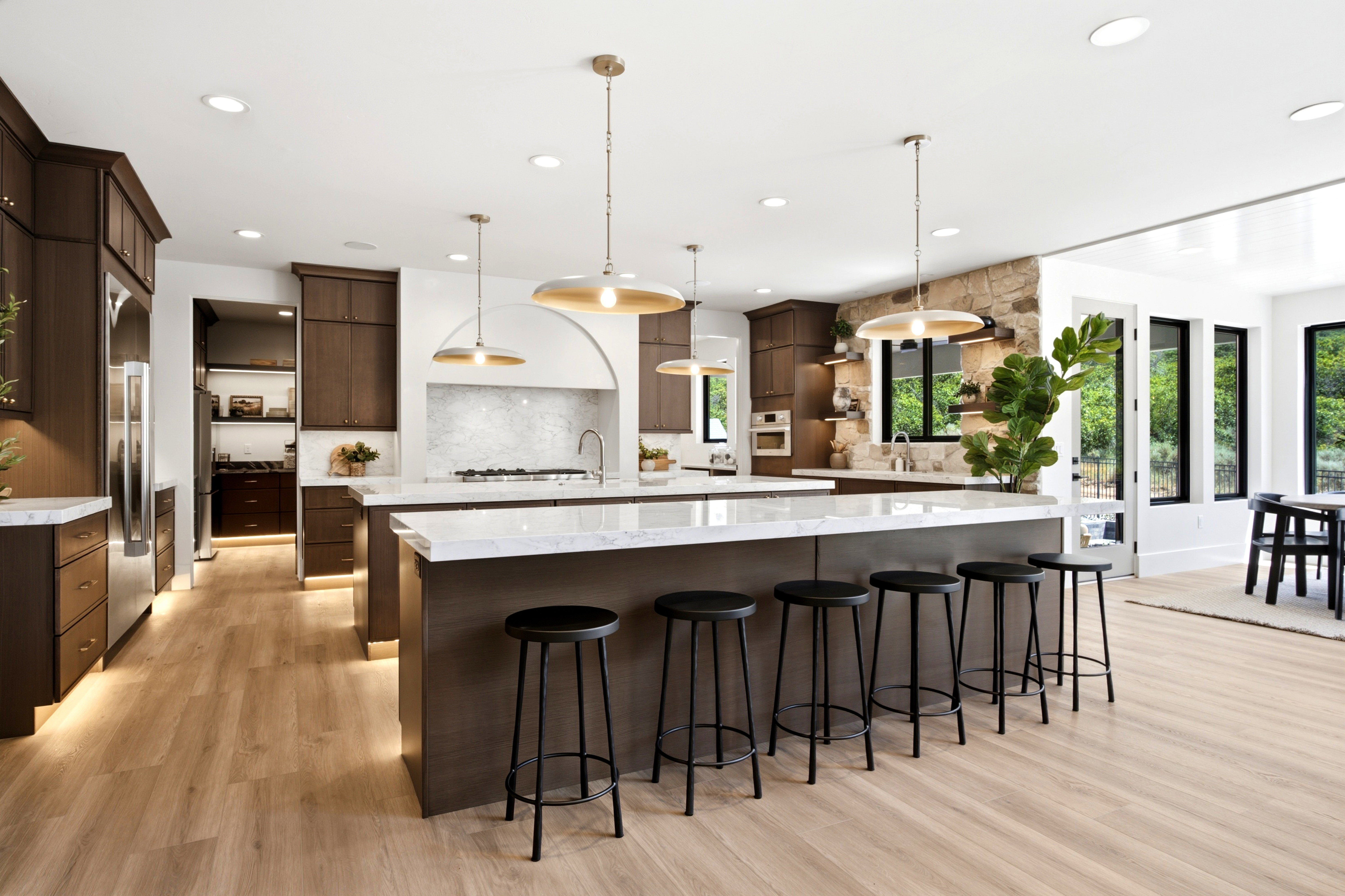 Gourmet kitchen featuring a large central island with seating, stainless steel appliances, dark wood cabinetry, light wood-finish flooring, and natural stone accent wall